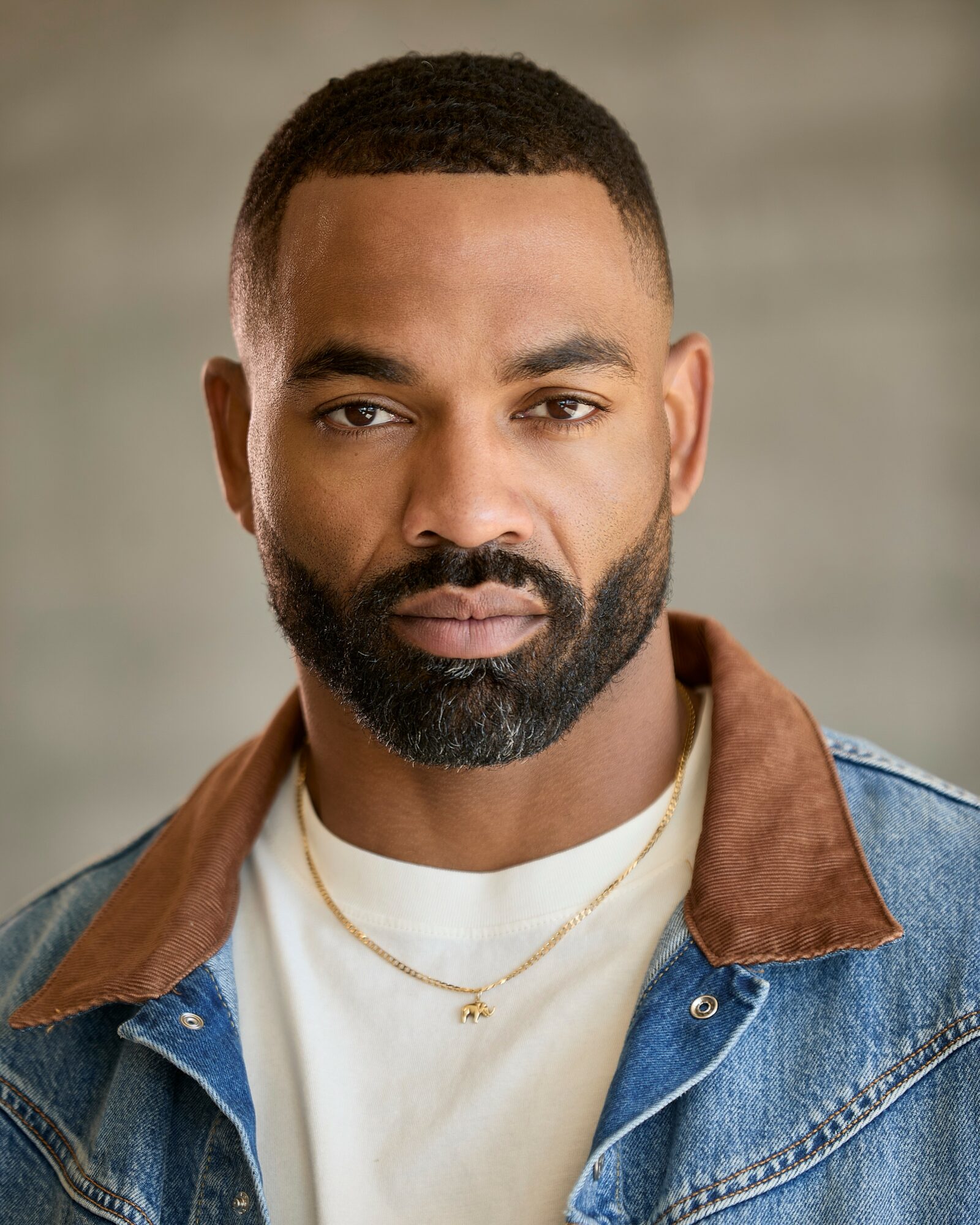 Portrait of a man with short hair and a beard, wearing a denim jacket with a brown collar and a white shirt, looking directly at the camera.