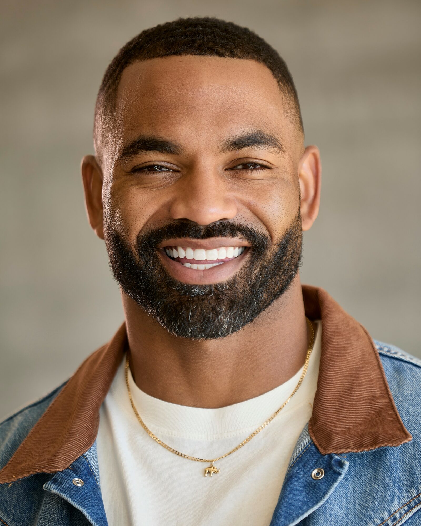 Smiling man with short hair and beard, wearing a denim jacket and gold necklaces, against a neutral background.