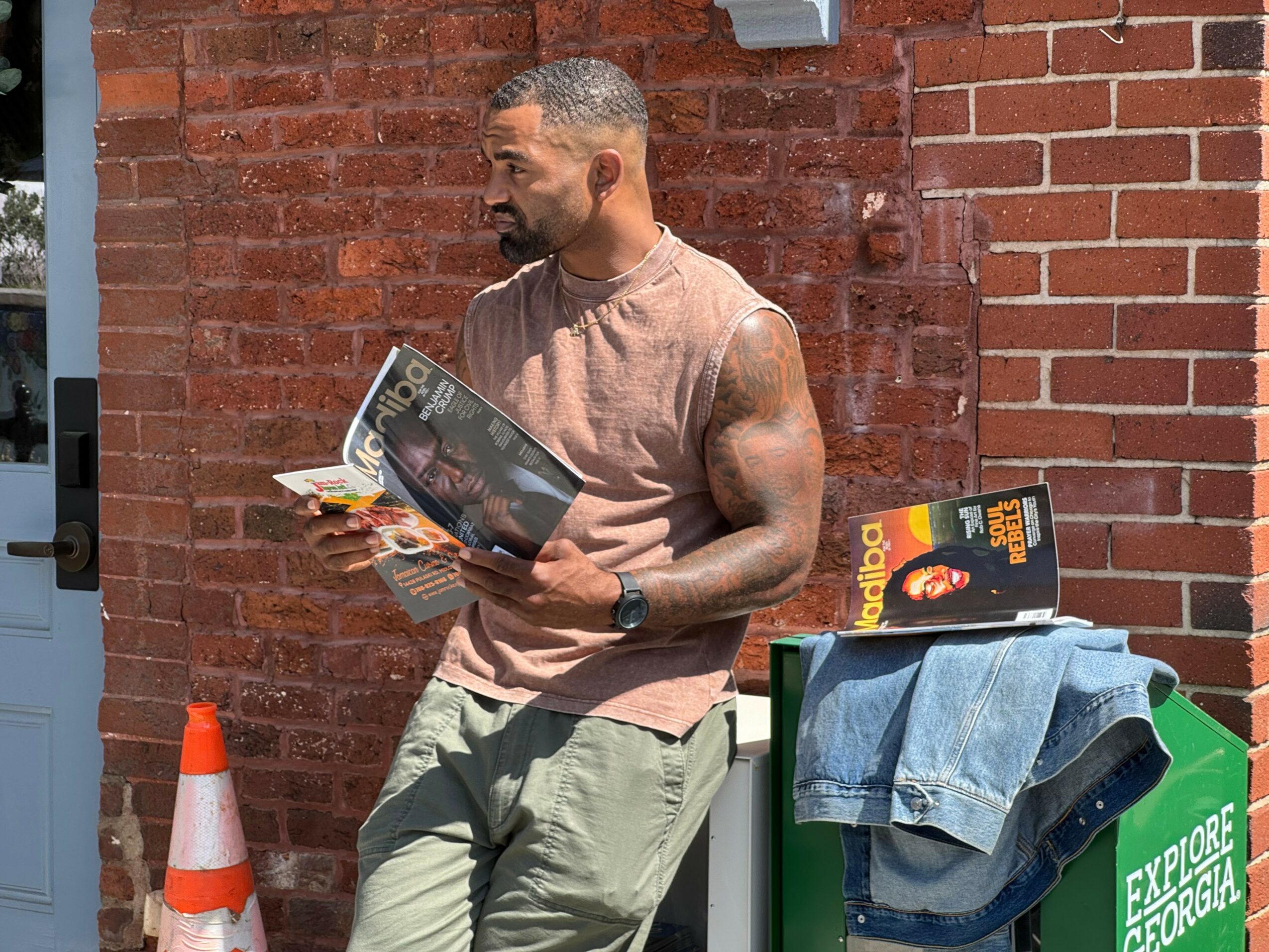 Man with tattoos reading a magazine outdoors against a brick wall, with a denim jacket on a green box.