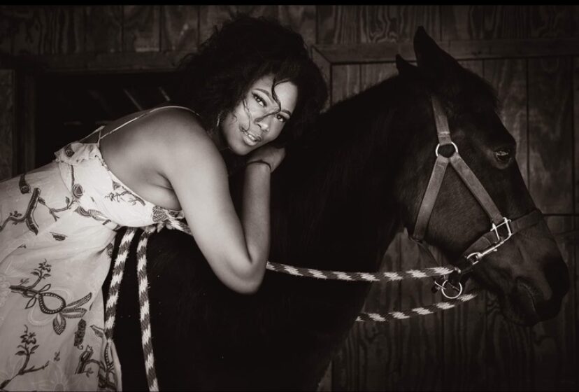 Woman with curly hair leaning on a horse, smiling, in a black and white photo, indoors with wooden background.