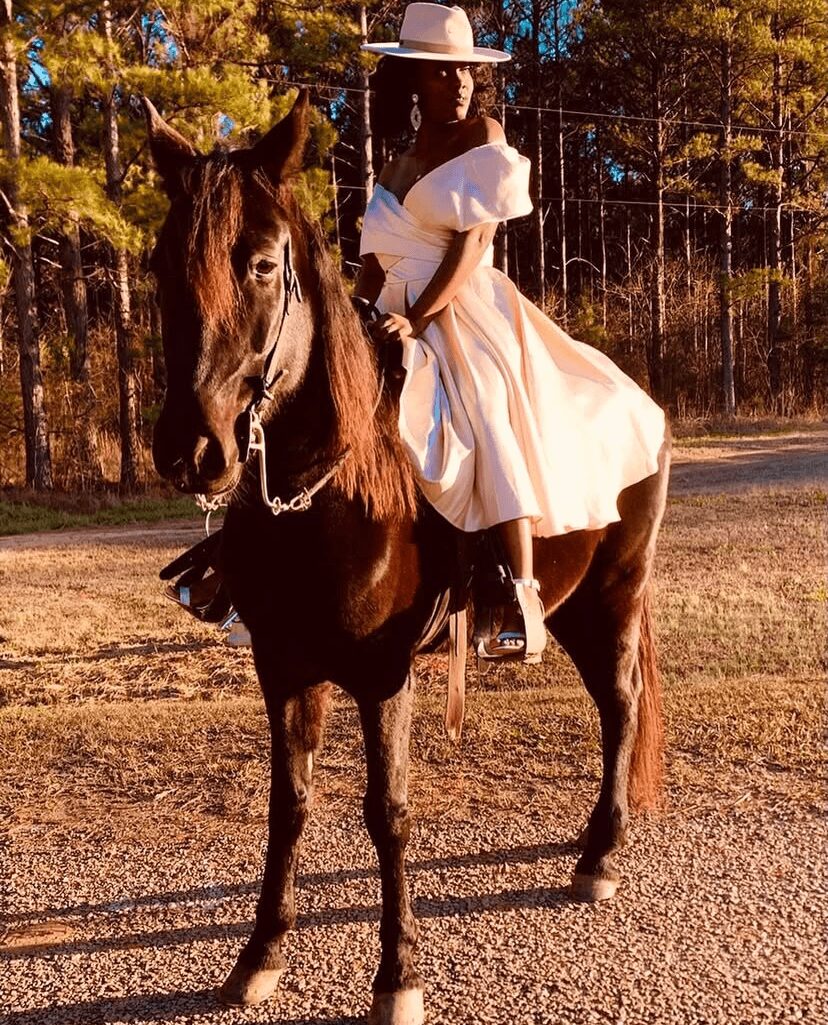 Person wearing a hat and white dress riding a dark horse outdoors with trees in background.