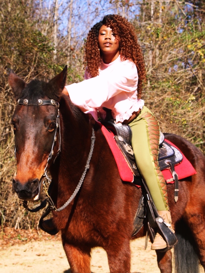 Young woman with curly hair riding a brown horse outdoors, surrounded by trees and foliage.