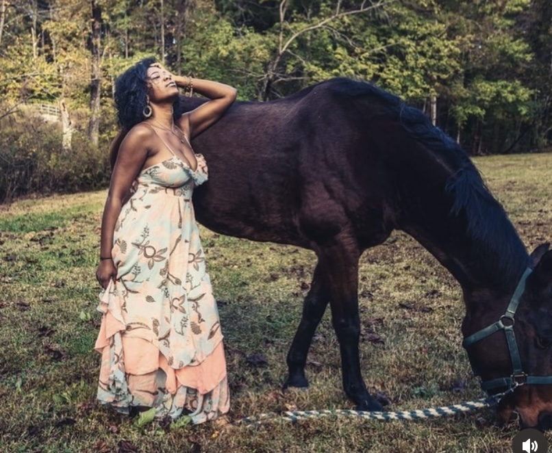 Woman in a floral dress stands outdoors with a horse, touching its neck, surrounded by trees and grass.