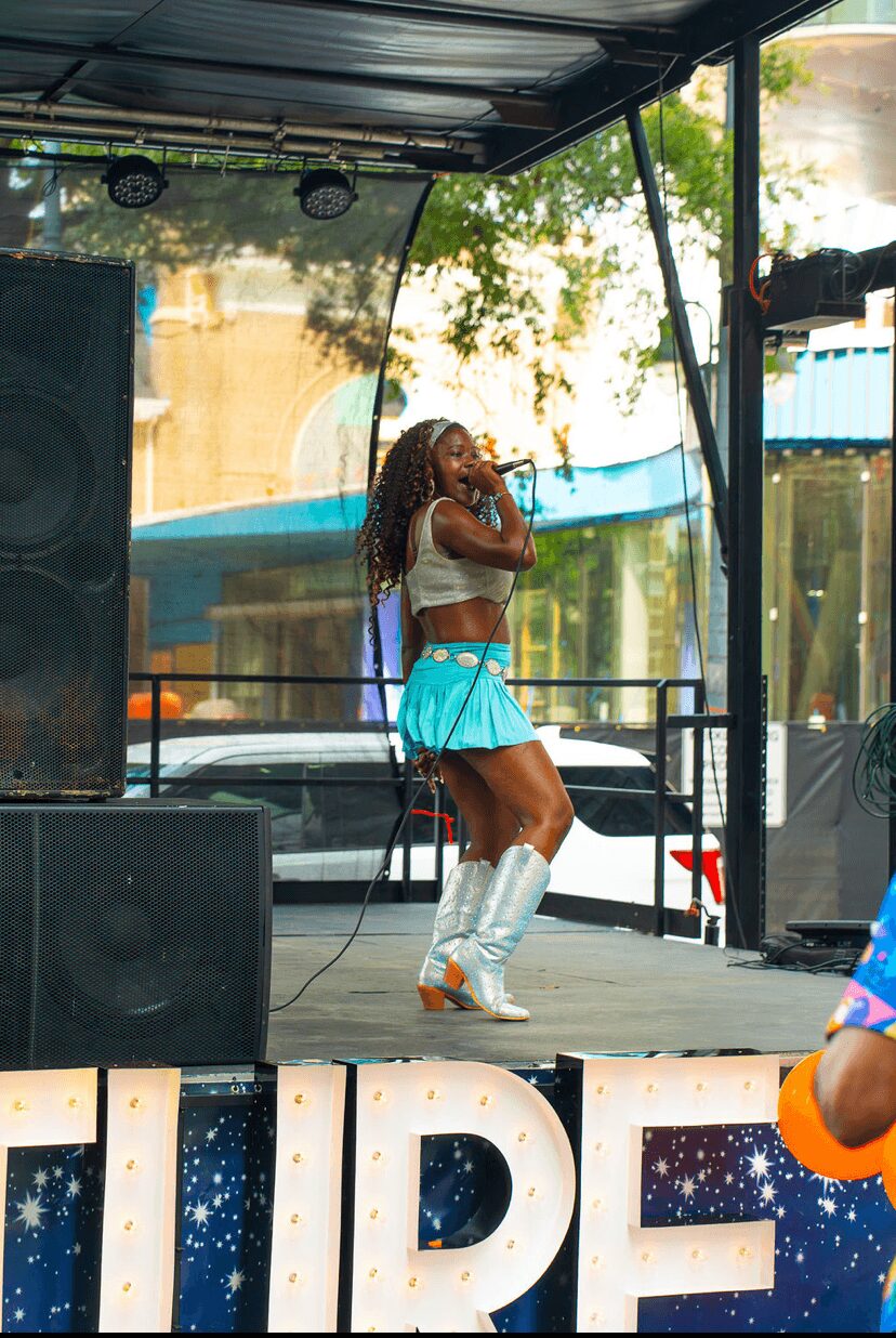 A woman with curly hair sings into a microphone on an outdoor stage, wearing a white top, turquoise skirt, and white boots.