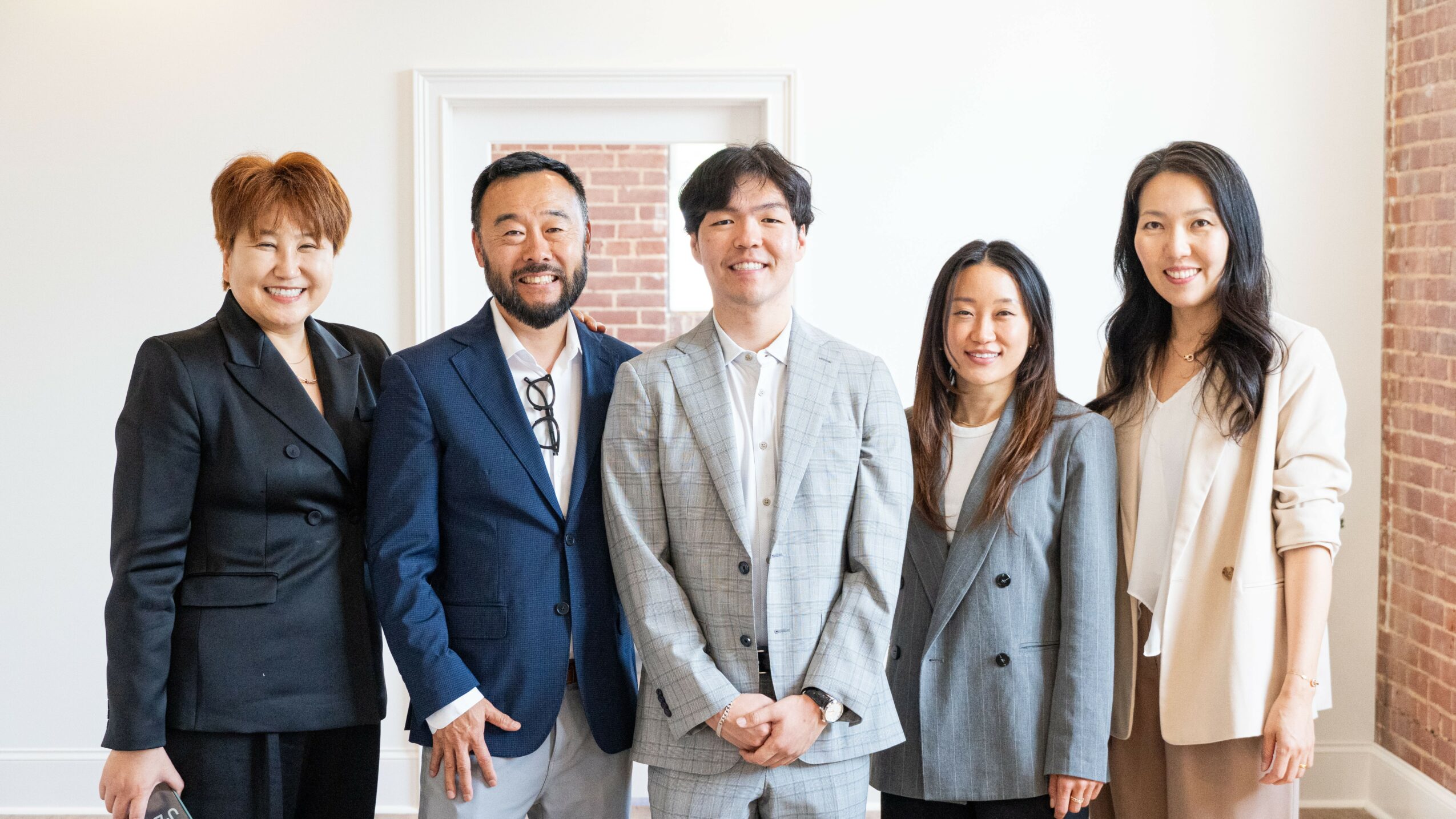 Five diverse professionals standing together indoors, smiling, with a white wall and brick wall in the background.