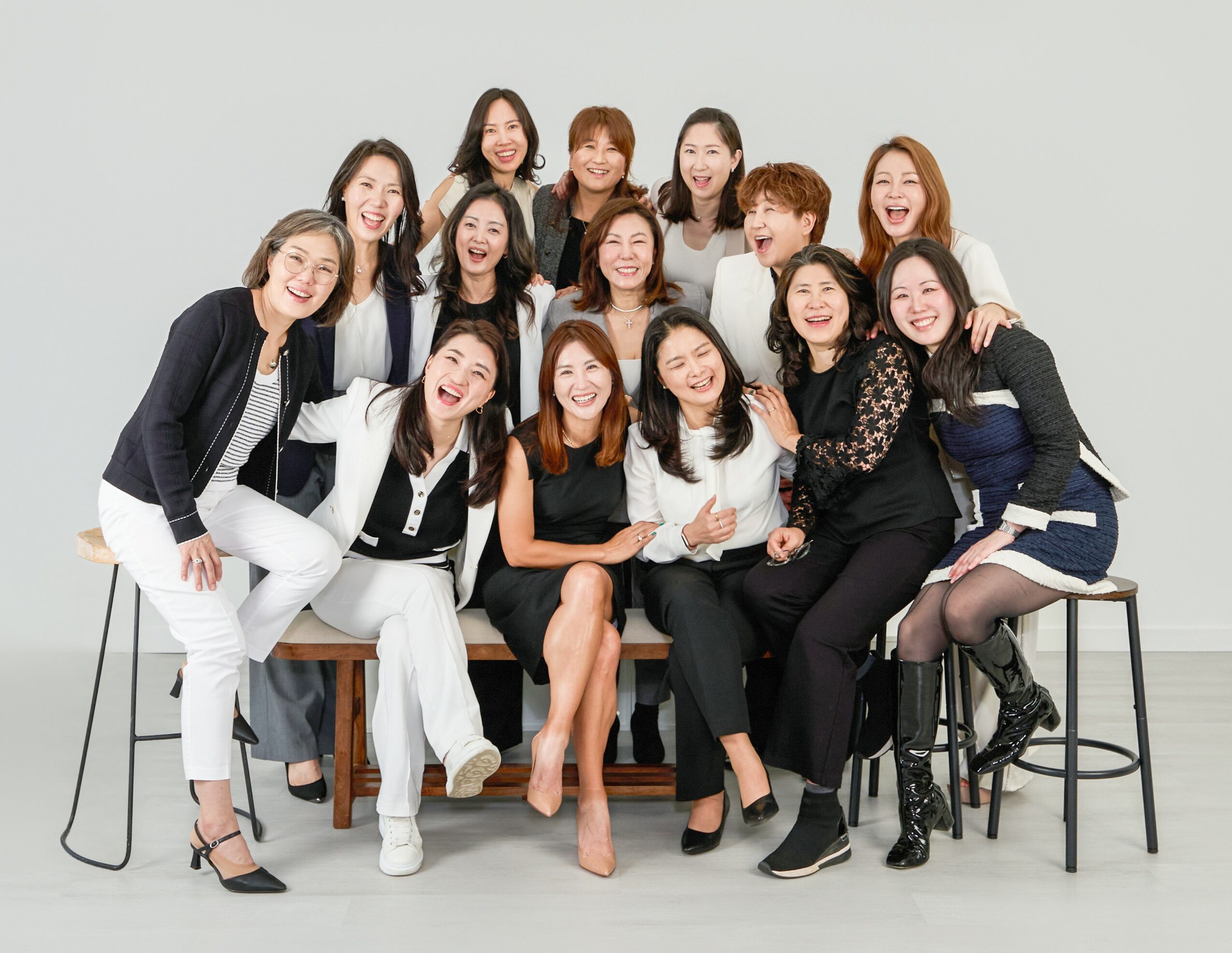 Group of fifteen women sitting and standing together, smiling, in a studio with a plain background.