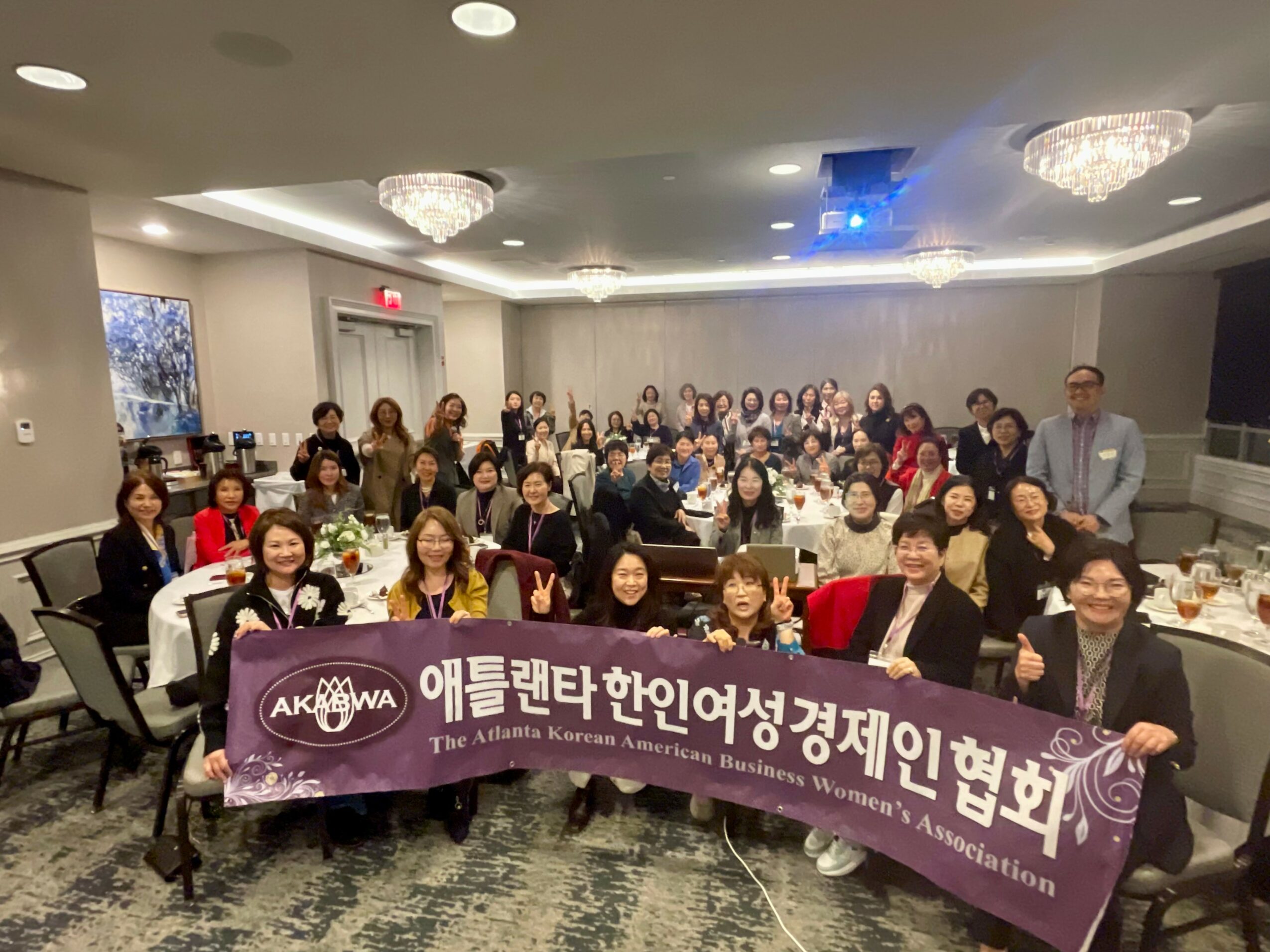 Group of people holding a banner in a banquet hall, with tables and chandeliers, facing forward and smiling.