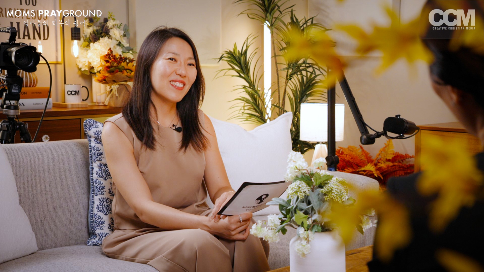 Woman sitting on a sofa, smiling, holding a tablet, surrounded by flowers and plants in a bright room.
