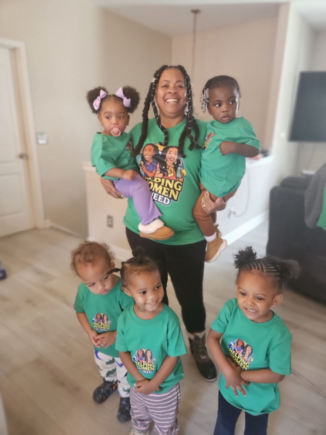 A woman holding two children, with three children standing in front, all wearing matching teal shirts, in a living room.