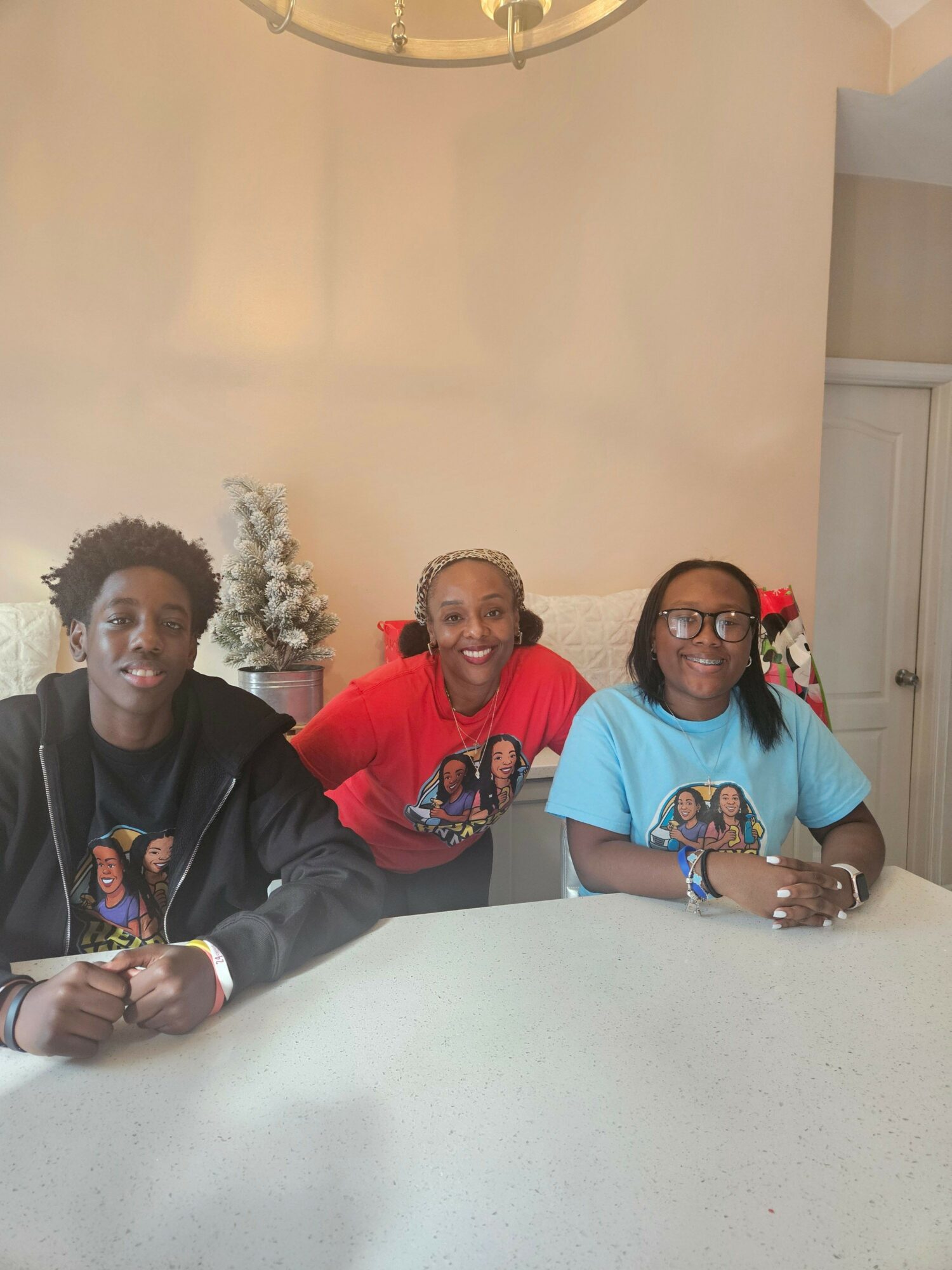 Three women sitting at a table, smiling, with a plain wall in the background.