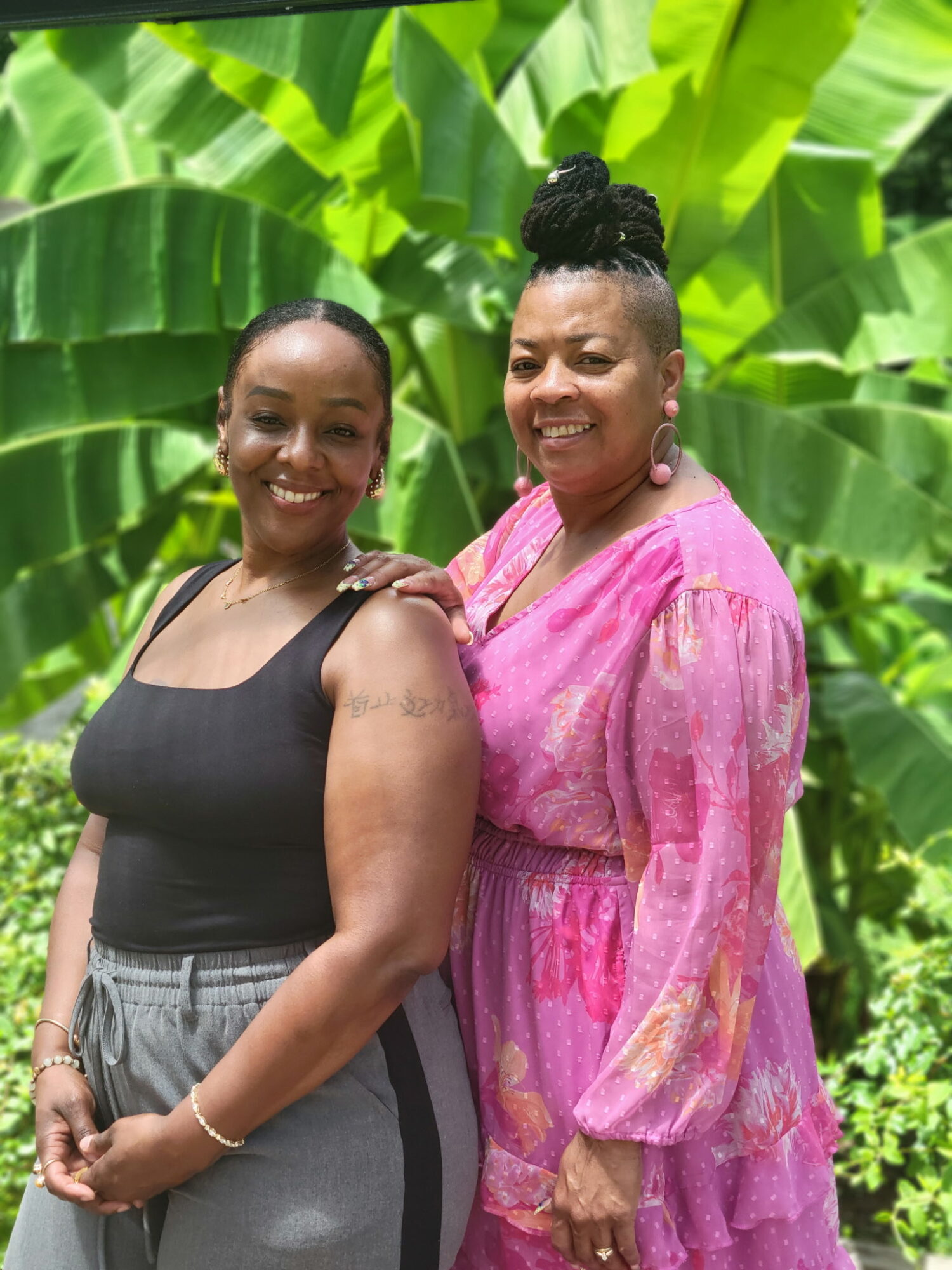 Two women smiling outdoors with large green leaves in the background, one in a black top and gray pants, the other in a pink dress.