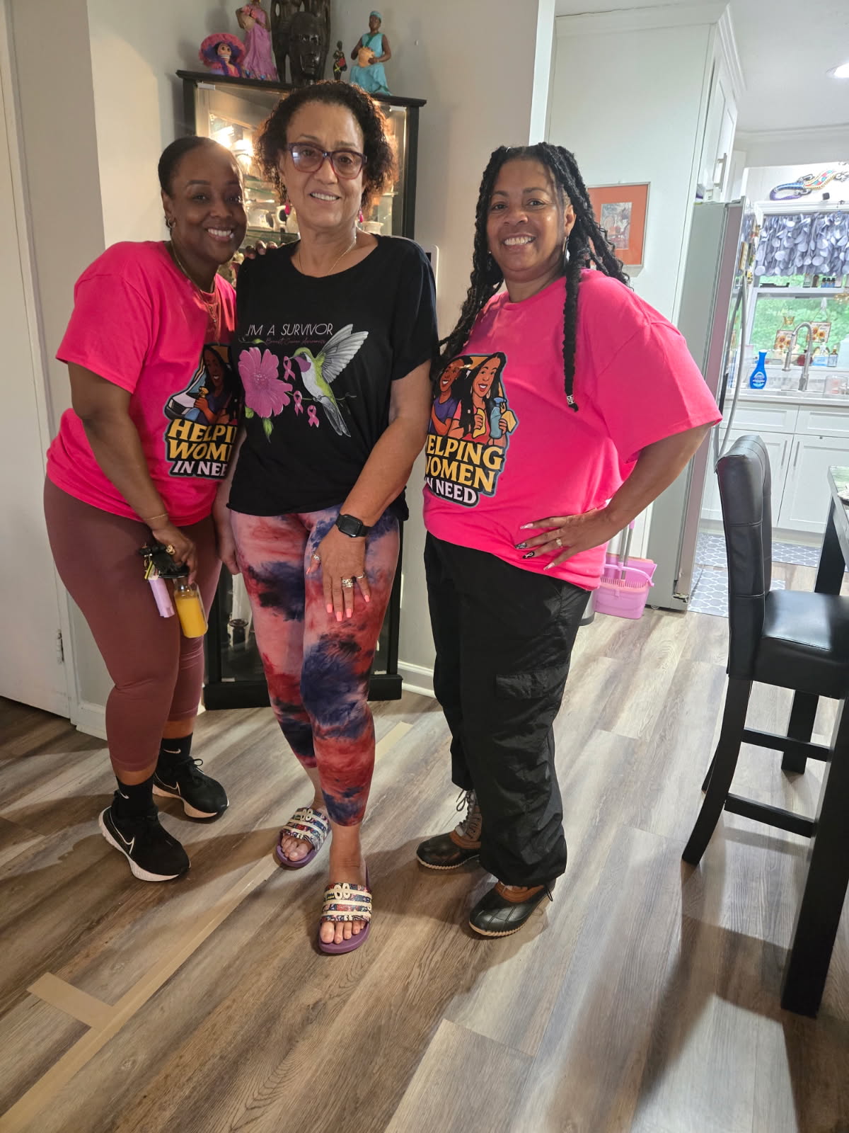 Three women standing indoors, smiling, with two wearing pink shirts and one in a black shirt, in a room with wooden floor.
