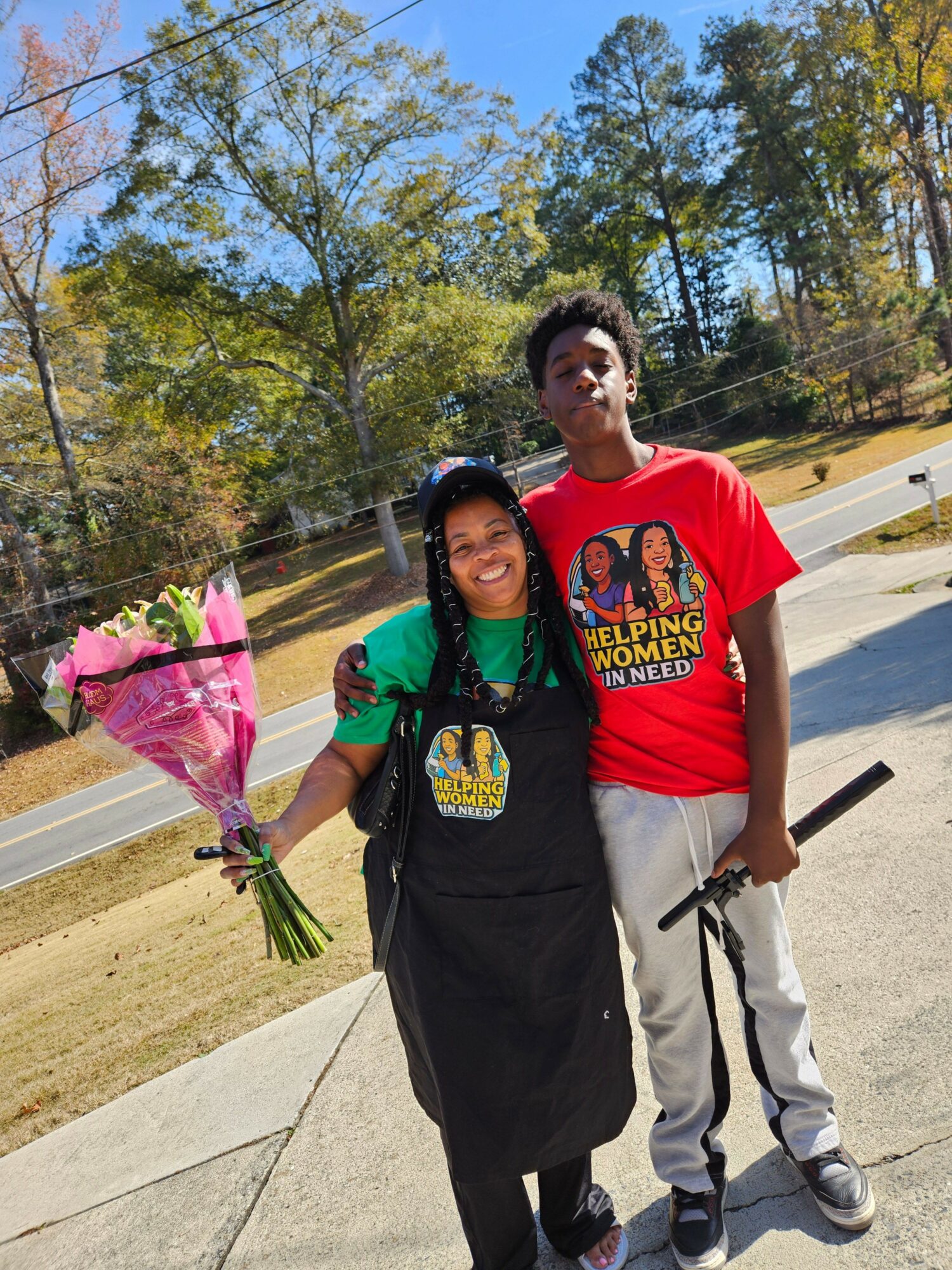 A young woman holding a bouquet of flowers stands next to a young man on a sidewalk, with trees and a road in the background.
