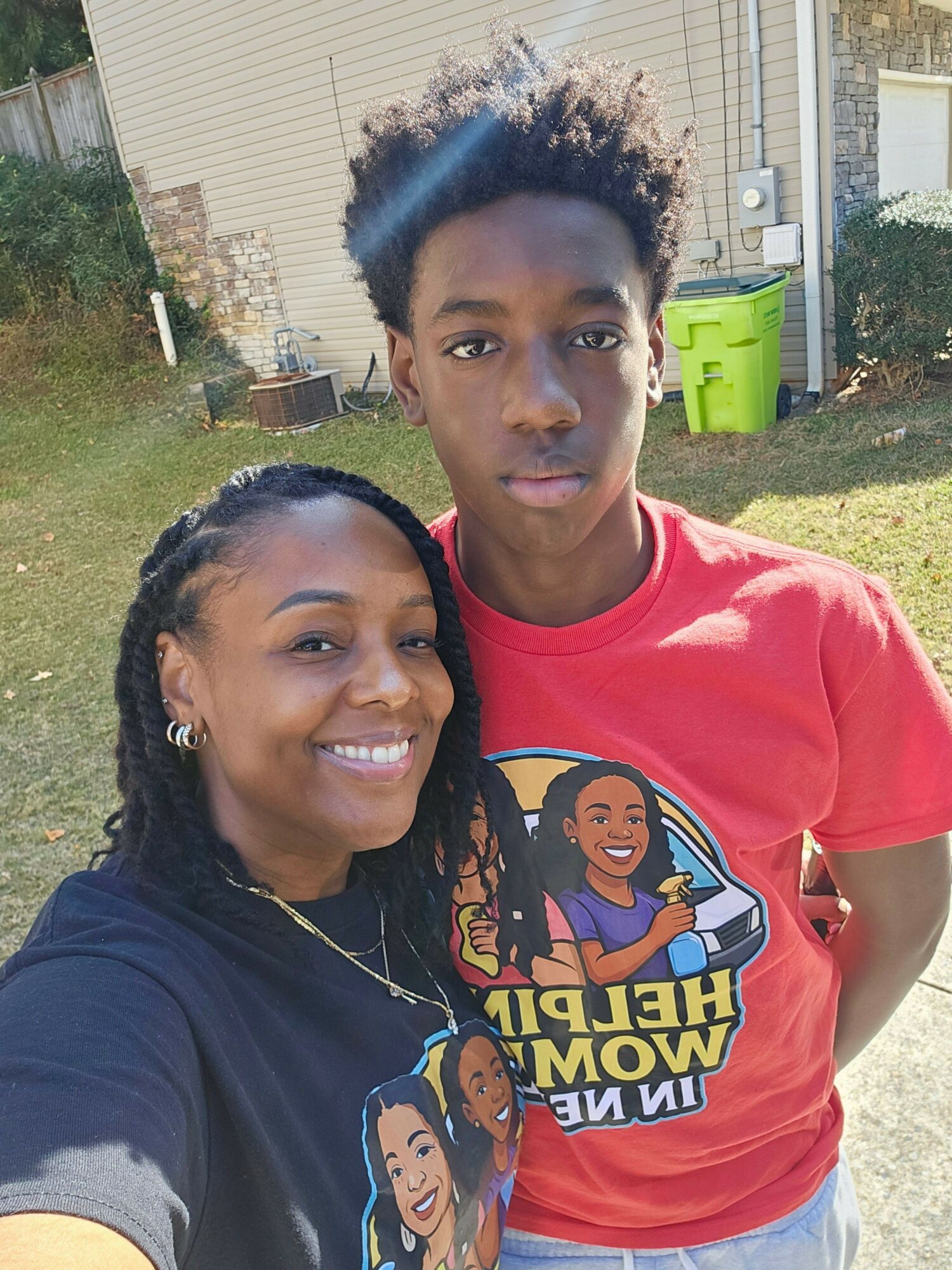 Two people smiling outdoors, one wearing a red shirt with a cartoon graphic, the other in a dark shirt, both looking at the camera.