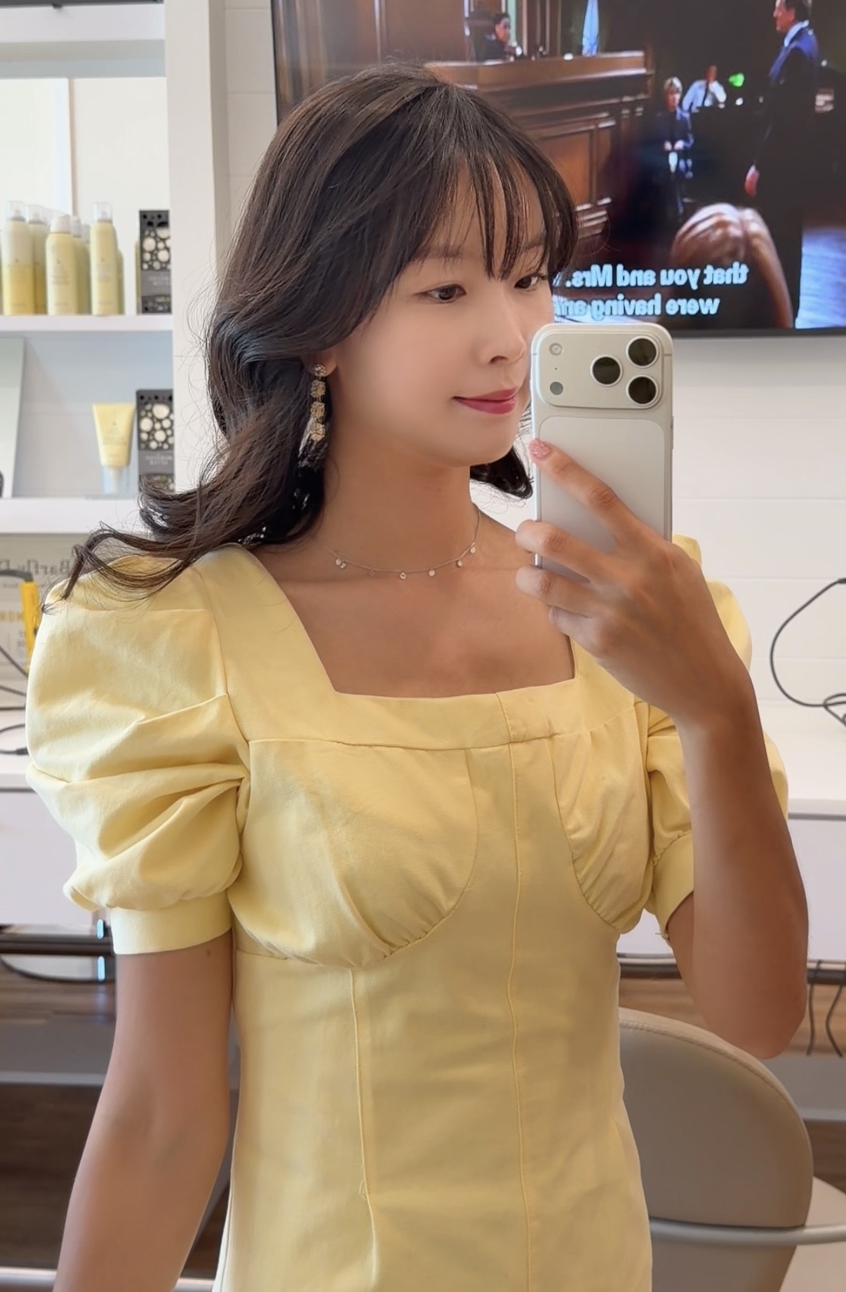 Woman with dark hair in a yellow dress taking a selfie in a mirror, background includes a TV and shelves.