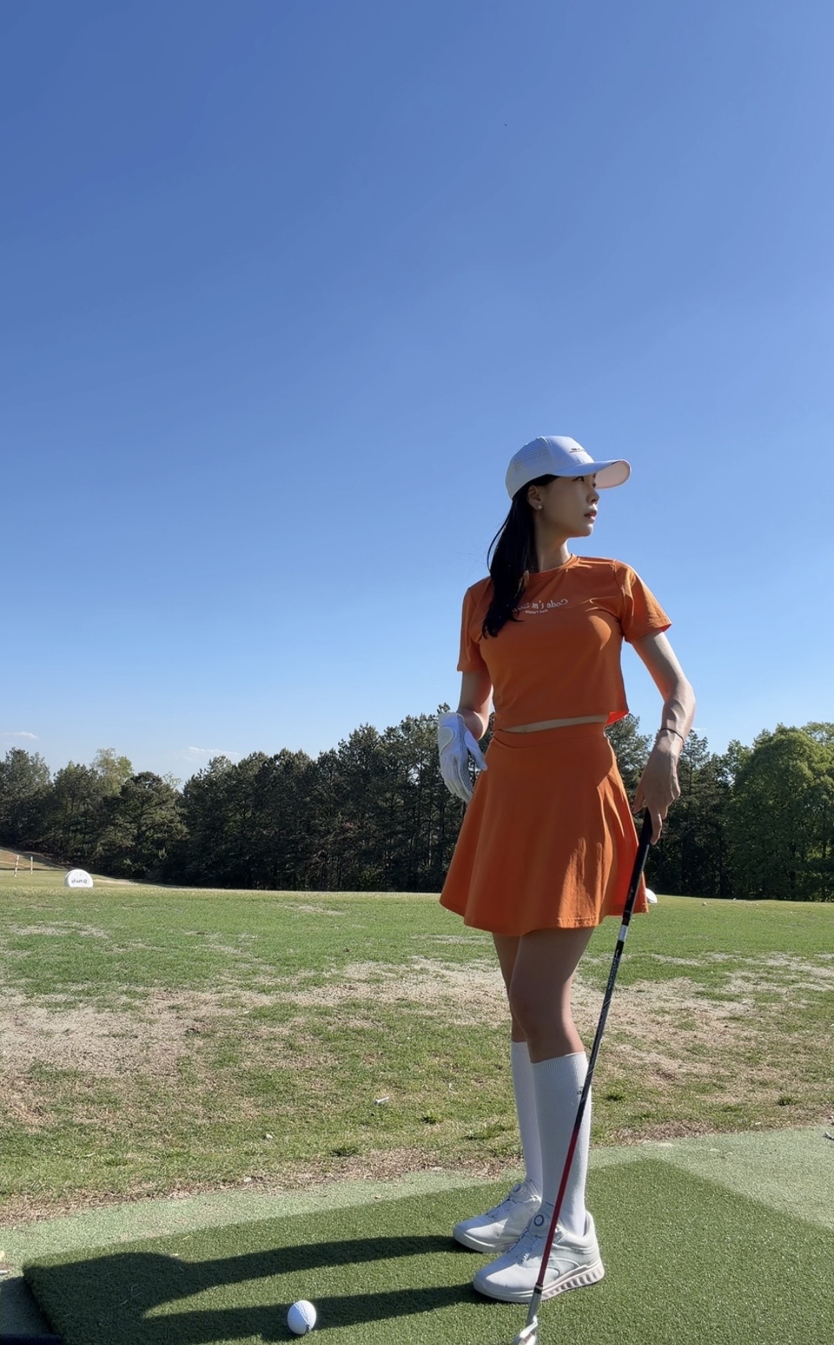 Woman in orange dress and white cap holding a golf club on a golf course, with trees and blue sky in background.
