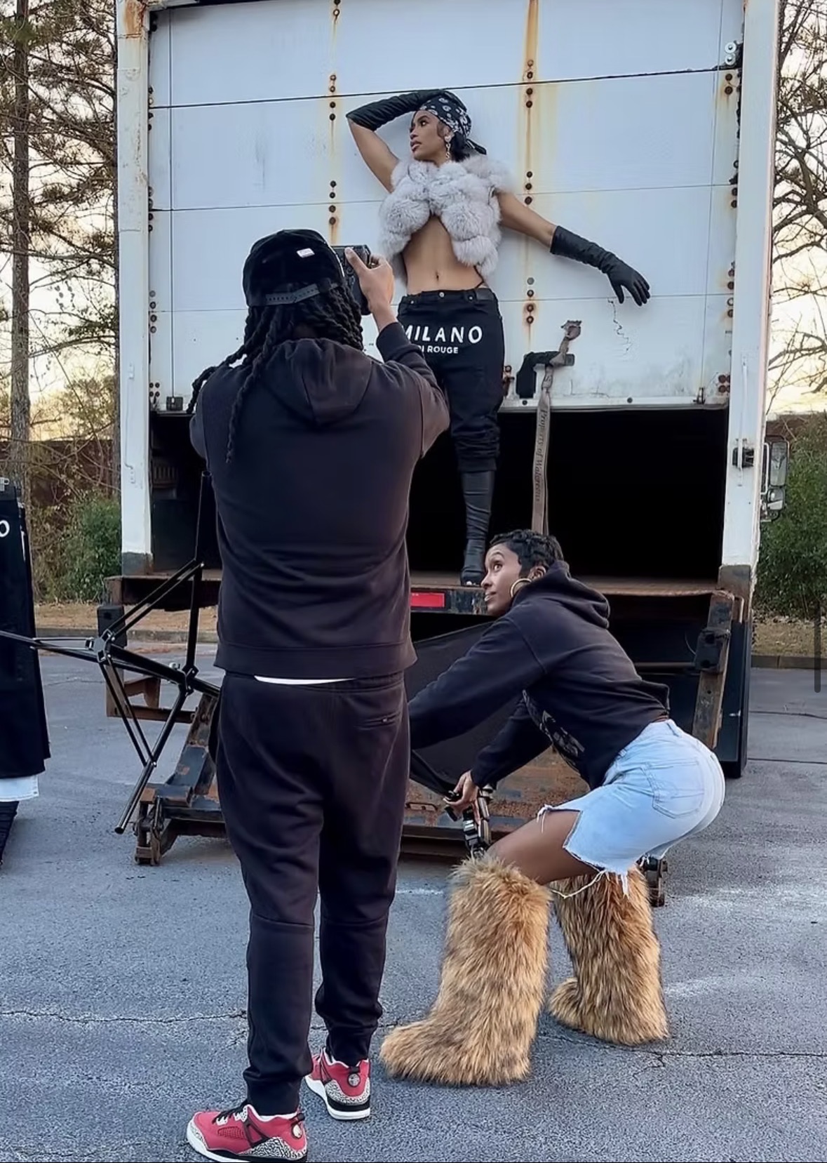 A woman in a stylish outfit poses on a truck, with two people in front taking photos, one crouching and one standing.