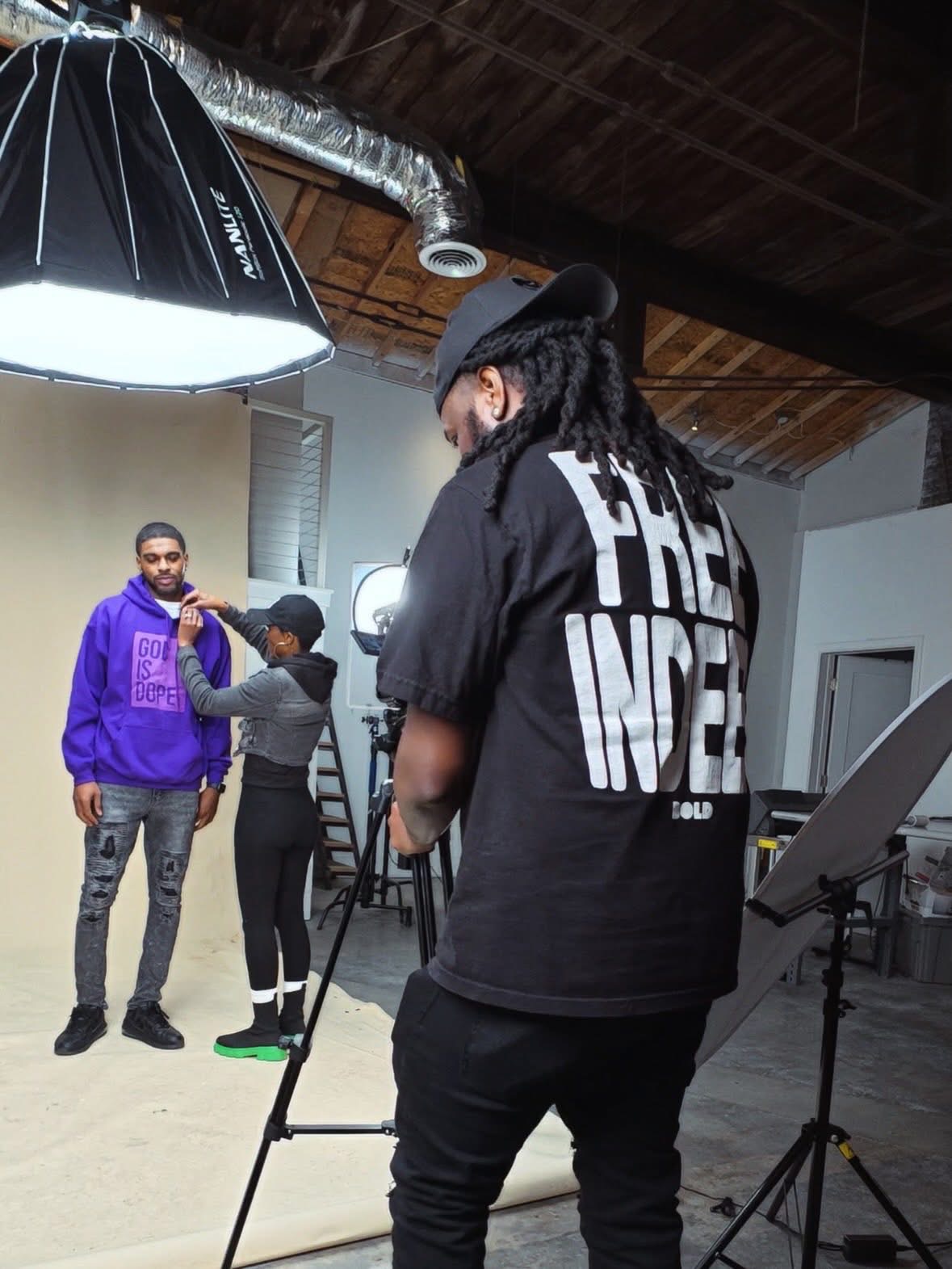 Three people in a studio with a large light, one person with dreadlocks and a hat, two others in casual clothing.