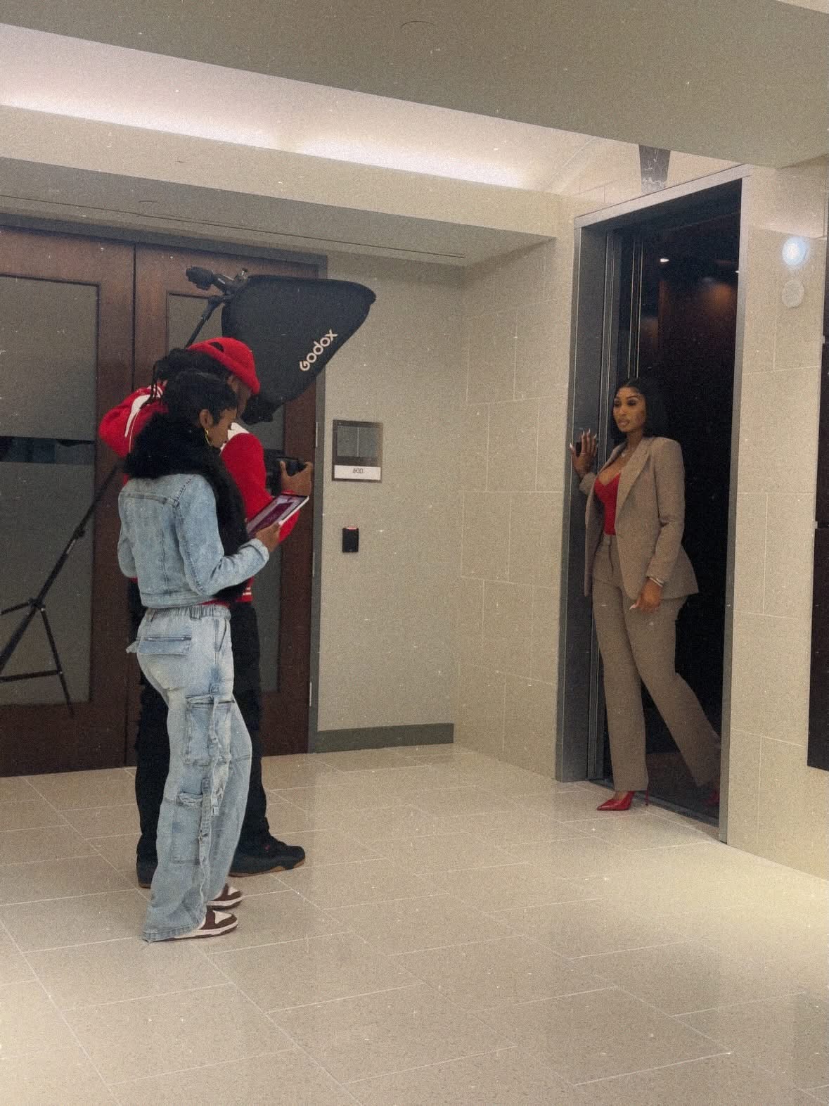 Three people standing in front of an open elevator door, one woman in a beige suit and two others in casual clothing, one with a red cap, reading a book or phone.