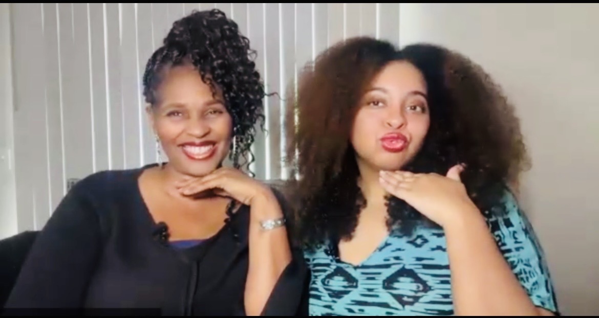 Two women sitting together, smiling, one with curly hair and the other with voluminous hair, in a room with vertical blinds.