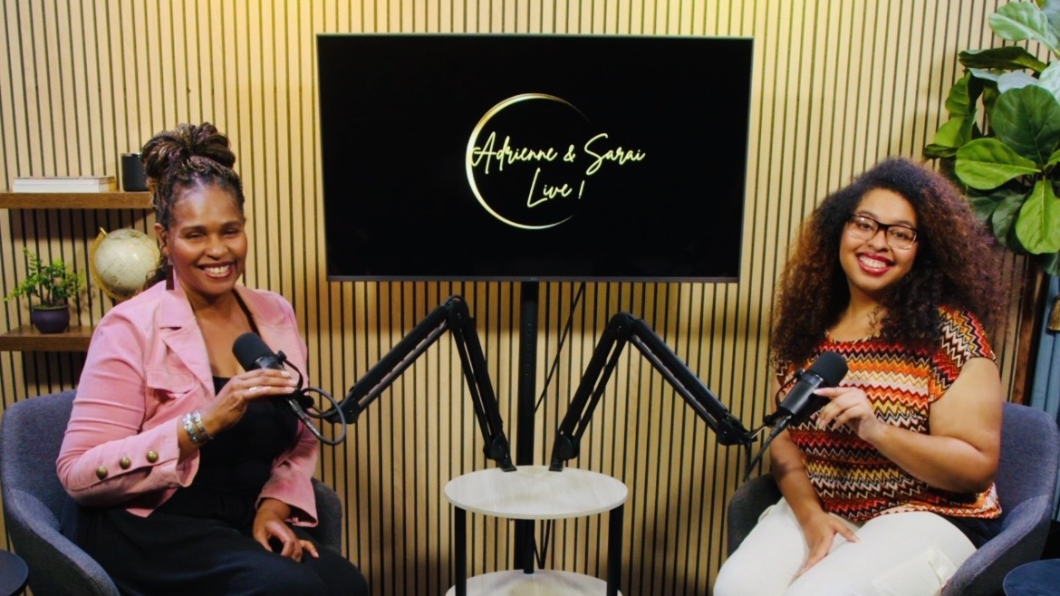 Two women sit in a recording studio with microphones, a screen displaying 'Adventures & Stories Live!', and a plant in the background.
