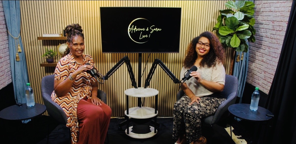 Two women sit in a studio with a screen behind them displaying text, holding microphones, with a small table and water bottles.