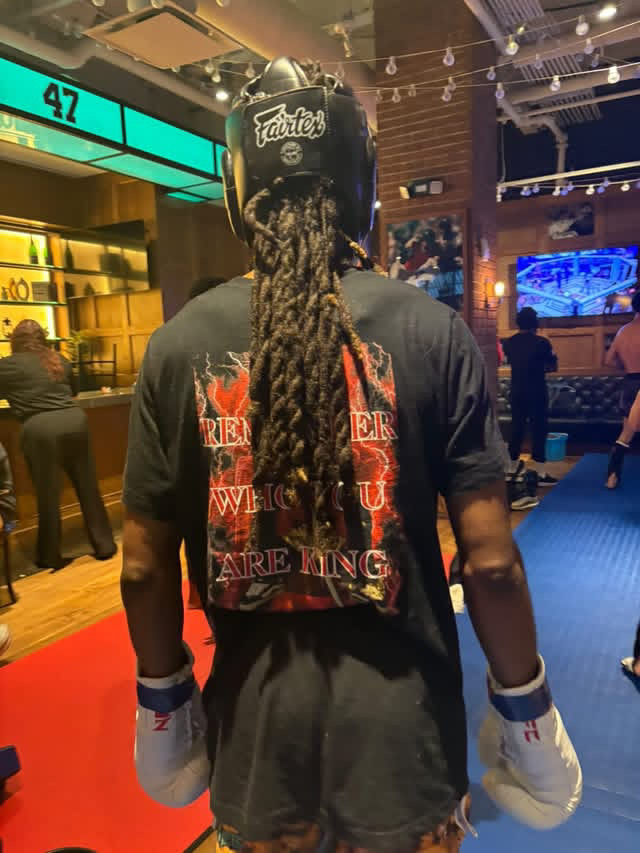 Person with long dreadlocks wearing a helmet and boxing gloves, standing in a gym or entertainment venue.