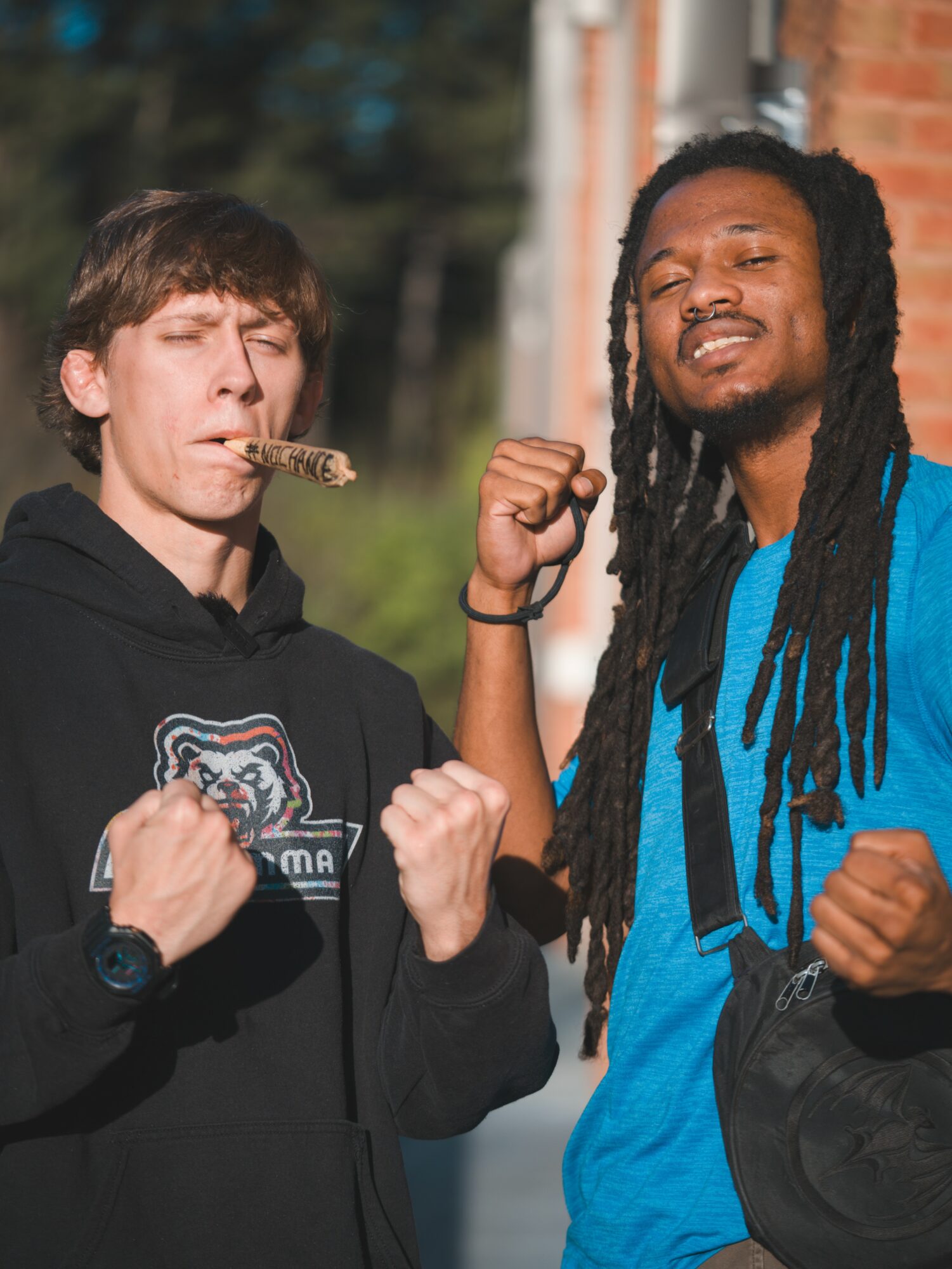 Two young men outdoors, one with a cookie in mouth and both showing fists, smiling confidently.