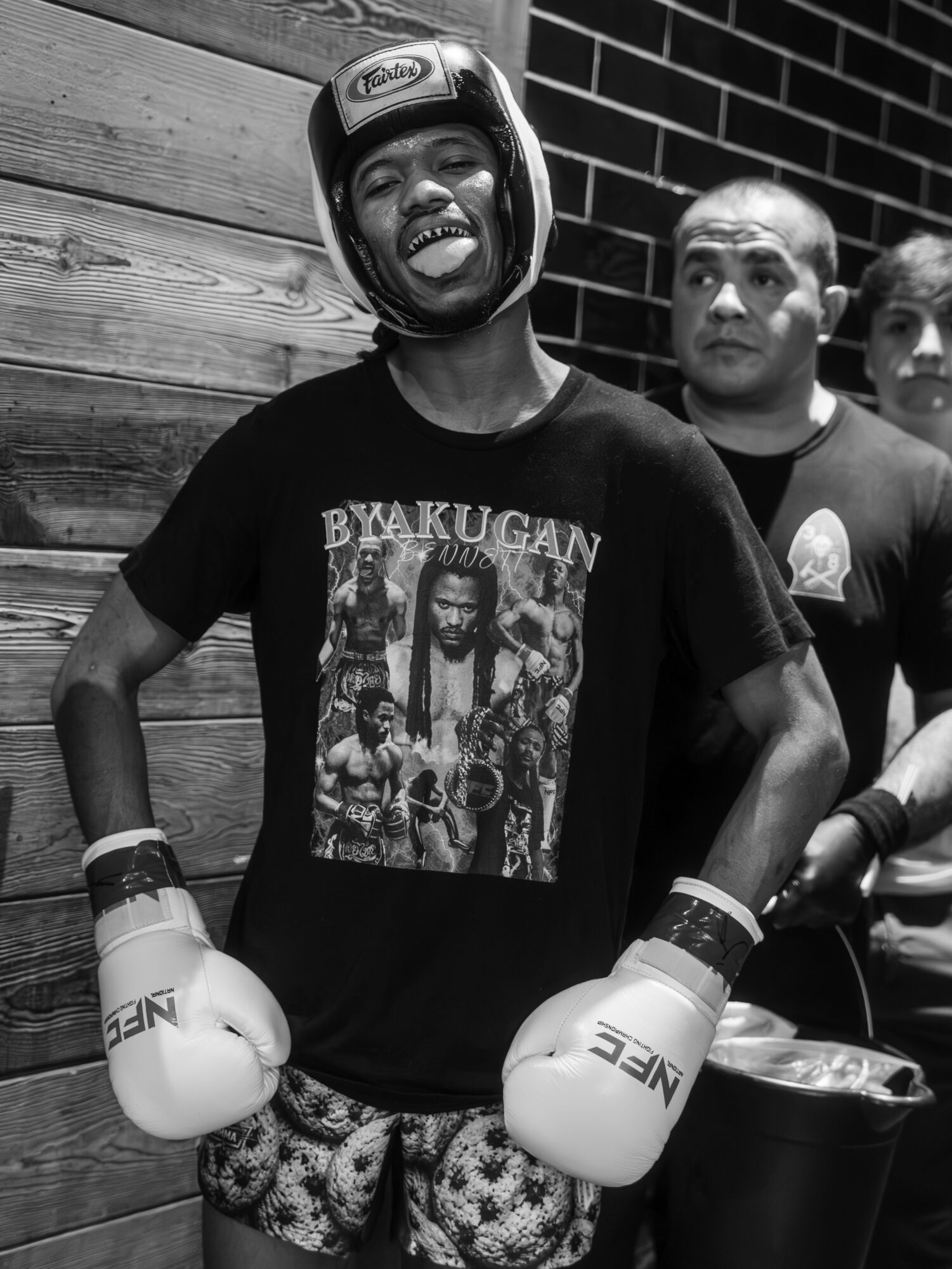 Person wearing boxing gloves and helmet, smiling, standing outdoors with a brick wall and other people in the background.