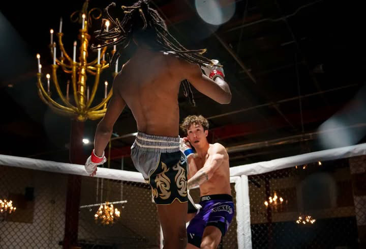 Mixed martial arts fight inside a cage with two fighters, one delivering a kick to the other, under chandeliers.