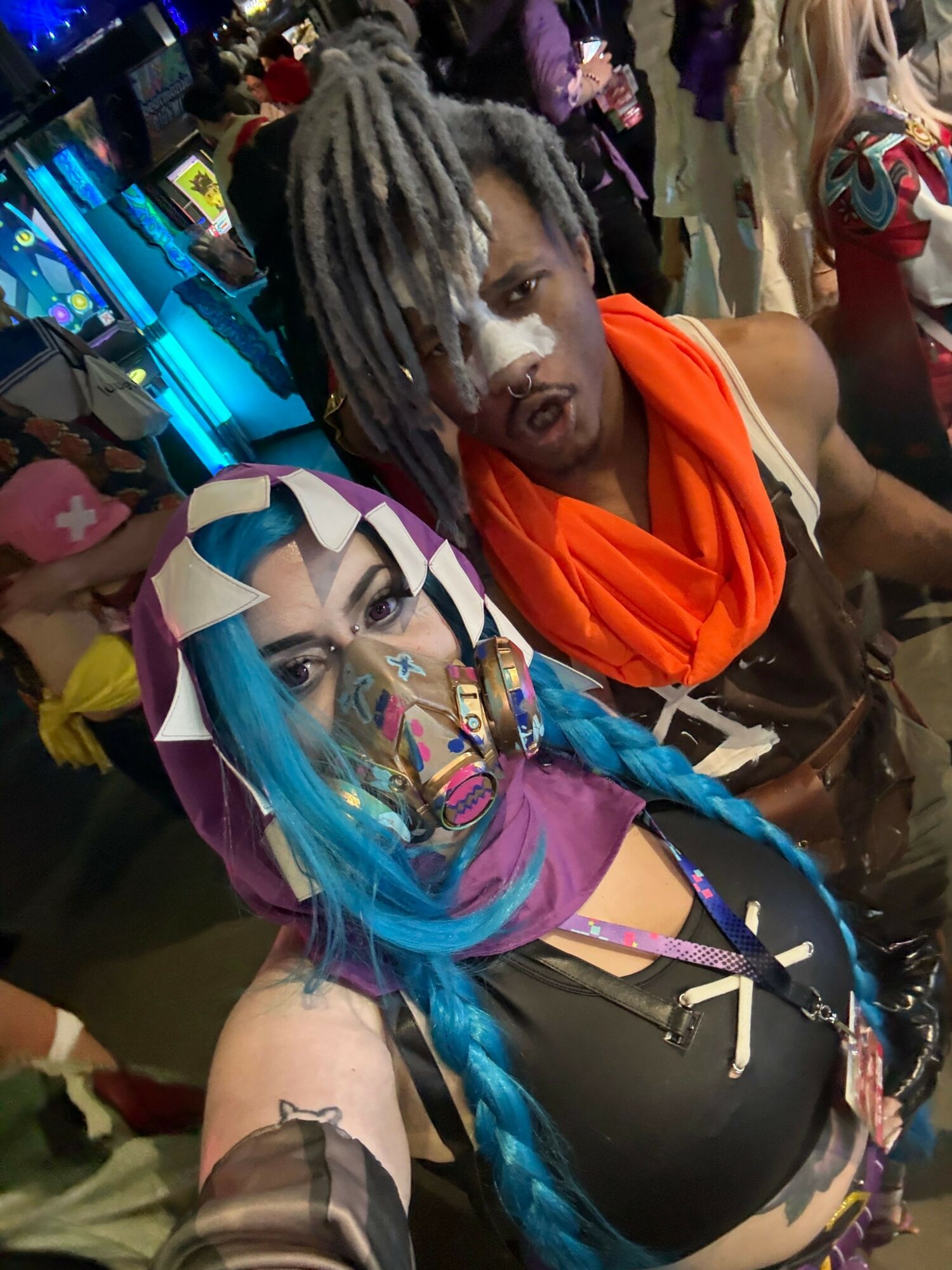 Two people lying down, one with gray dreadlocks and orange clothing, the other with colorful hair and accessories, in a crowded setting.