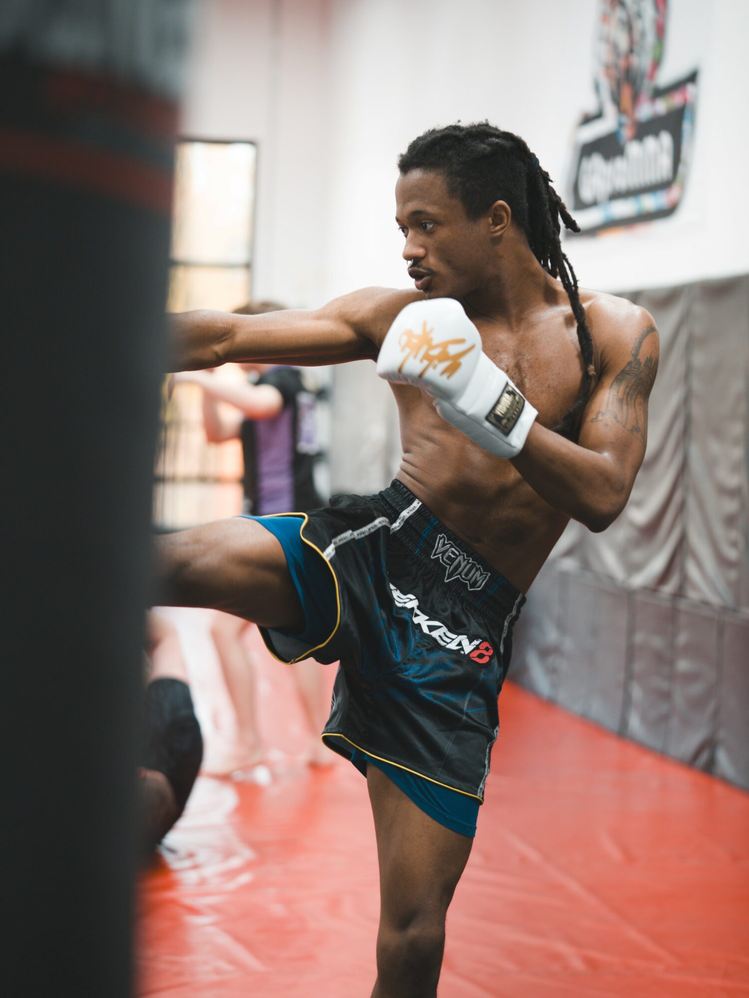 Male martial artist practicing kick in gym, wearing gloves and shorts, with a focused expression.