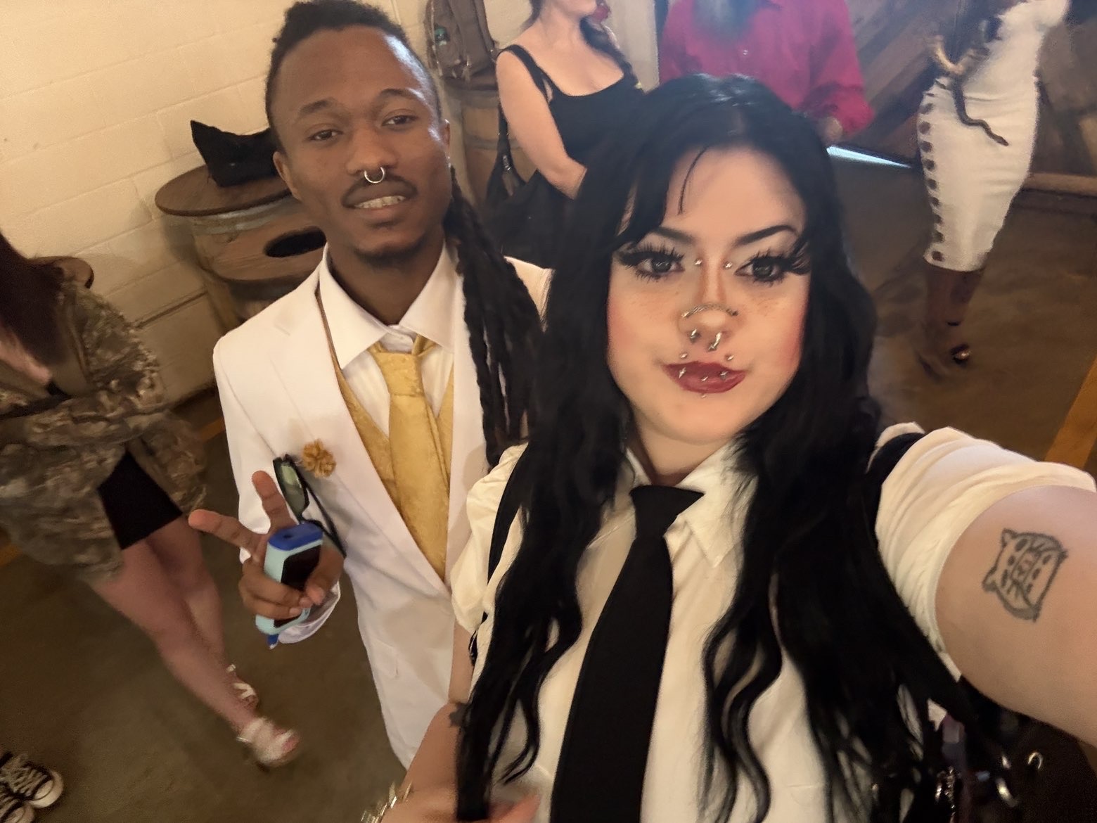 Two people taking a selfie at a social event, with others in the background, in a room with wooden floors.