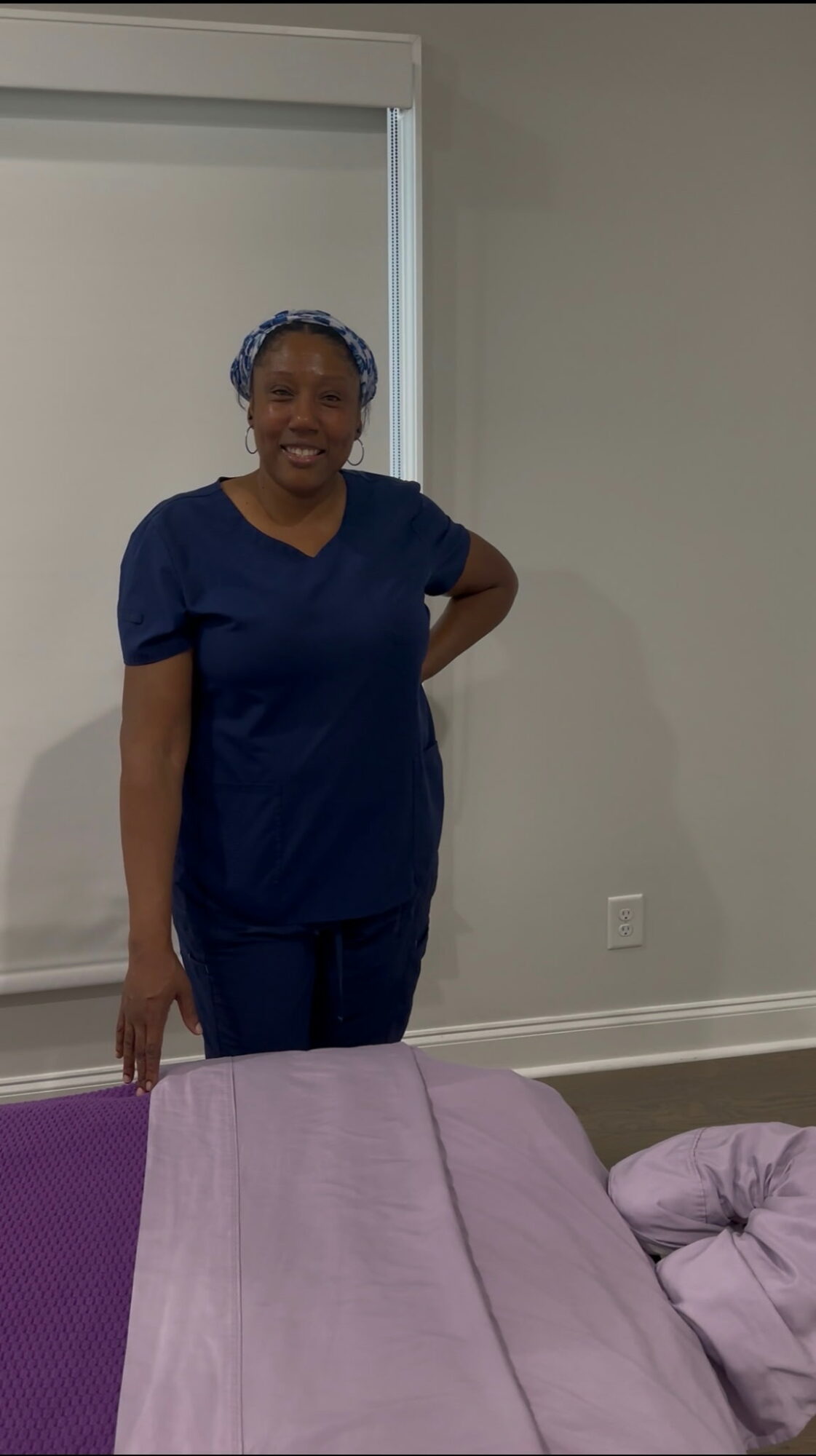 Woman standing indoors, smiling, wearing navy scrubs and a head covering, with a bed in the foreground.