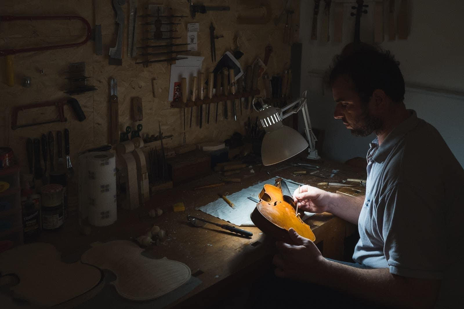 Person working on a guitar in a workshop with tools on the wall, illuminated by a desk lamp.