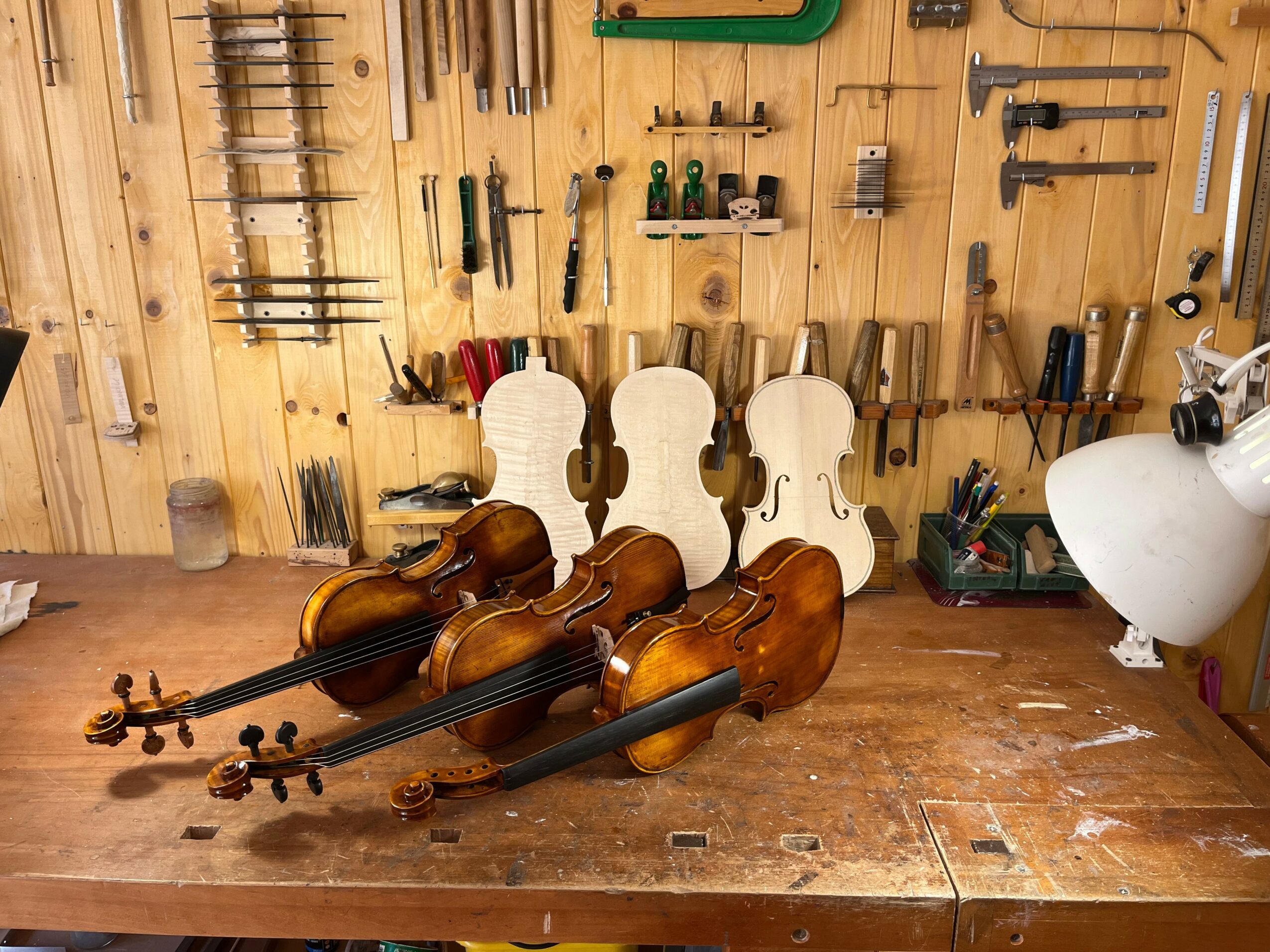 Three violins with bows rest on a wooden workbench in a workshop with tools on the wall behind.