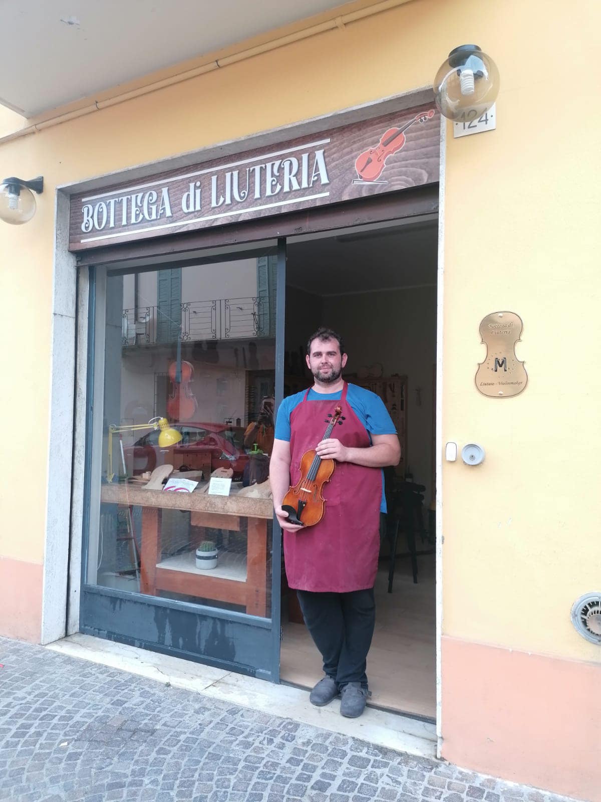 Man standing in doorway holding a guitar, wearing a red apron and blue shirt, outside a jewelry store.