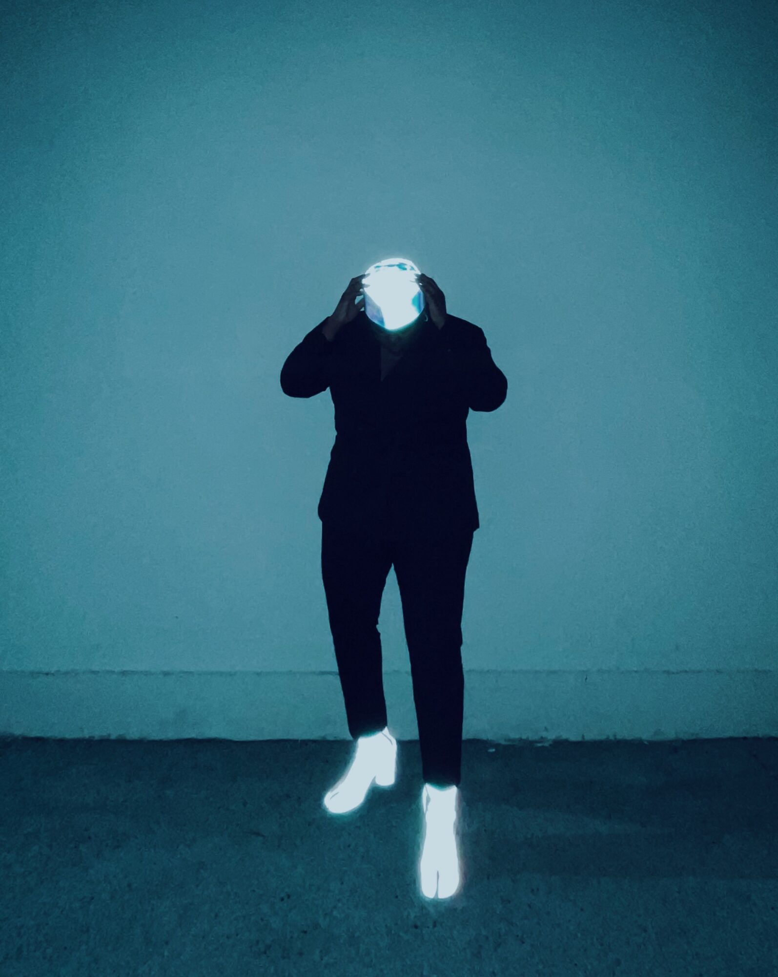 Person standing against a plain wall, holding a mask or object over their face, with bright white shoes and dark clothing.