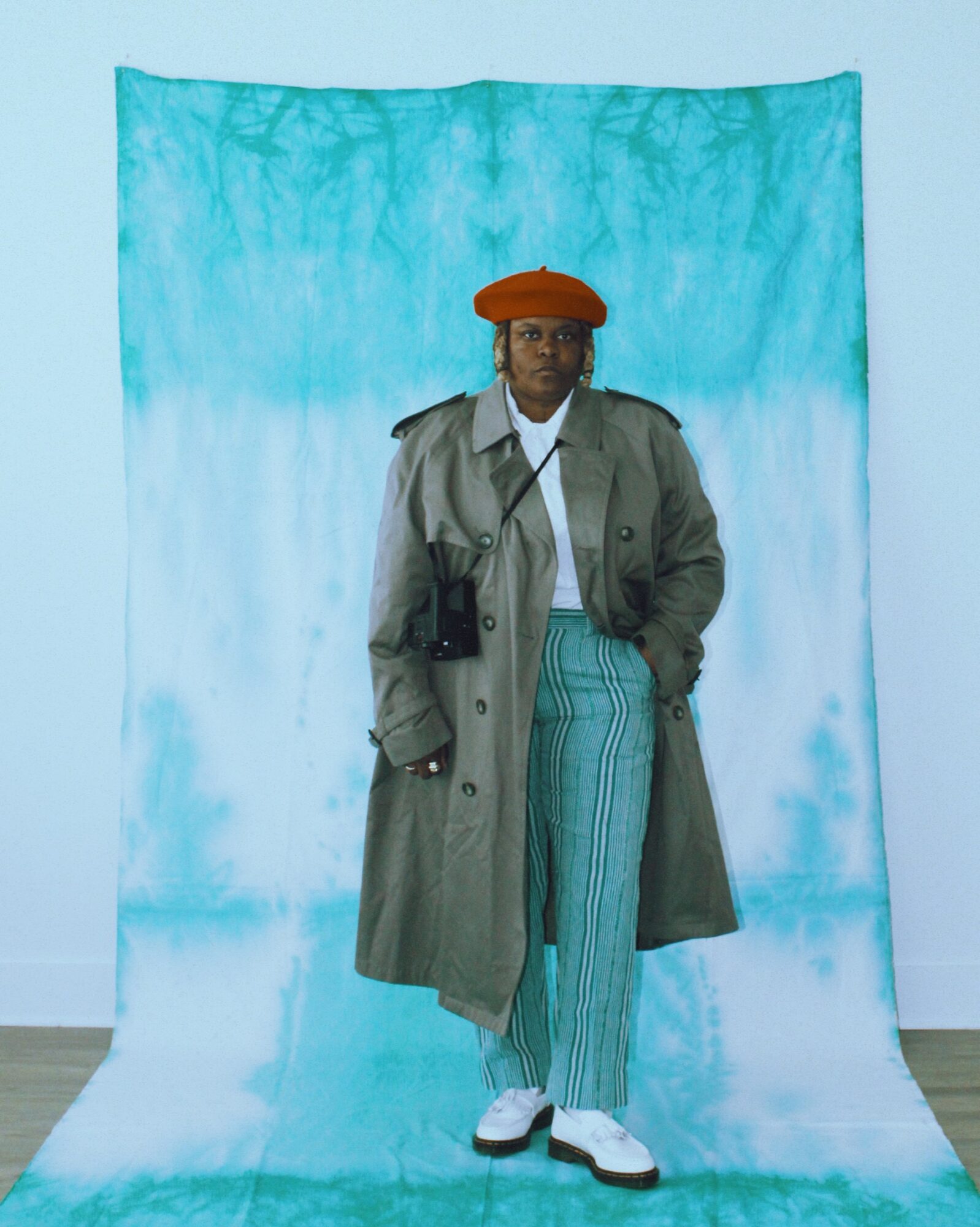 Person standing in front of a blue backdrop, wearing a trench coat, striped pants, white shoes, and an orange beret.