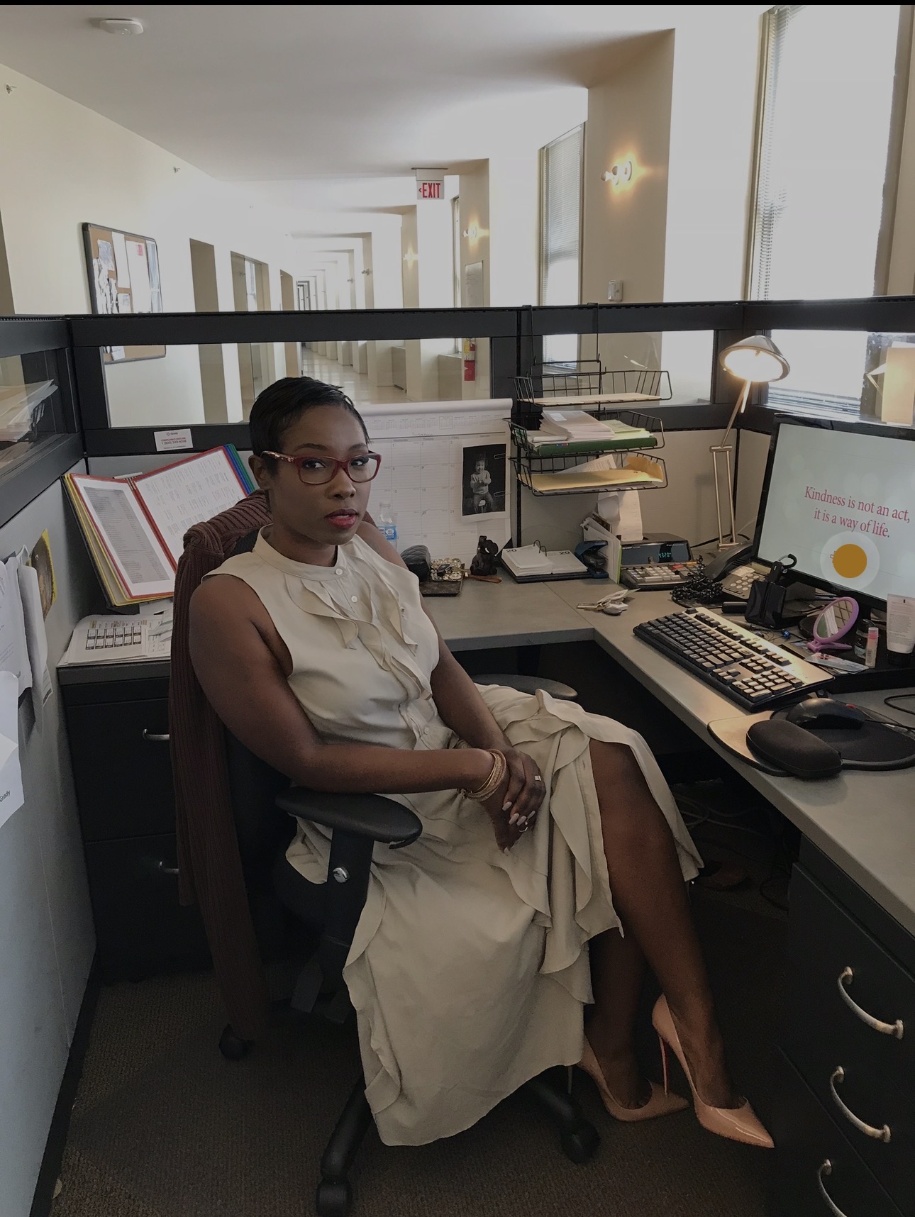 Woman sitting at a desk in an office, surrounded by computers, books, and office supplies, with windows in the background.
