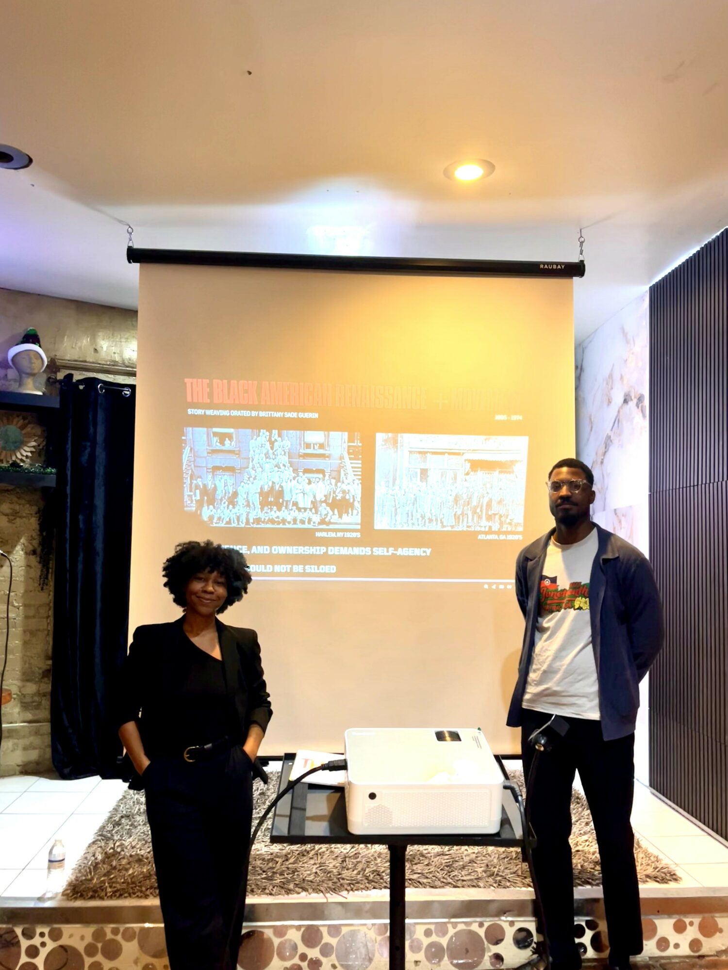 Two people standing in front of a projection screen in a room, one woman and one man, with a projector on a table.