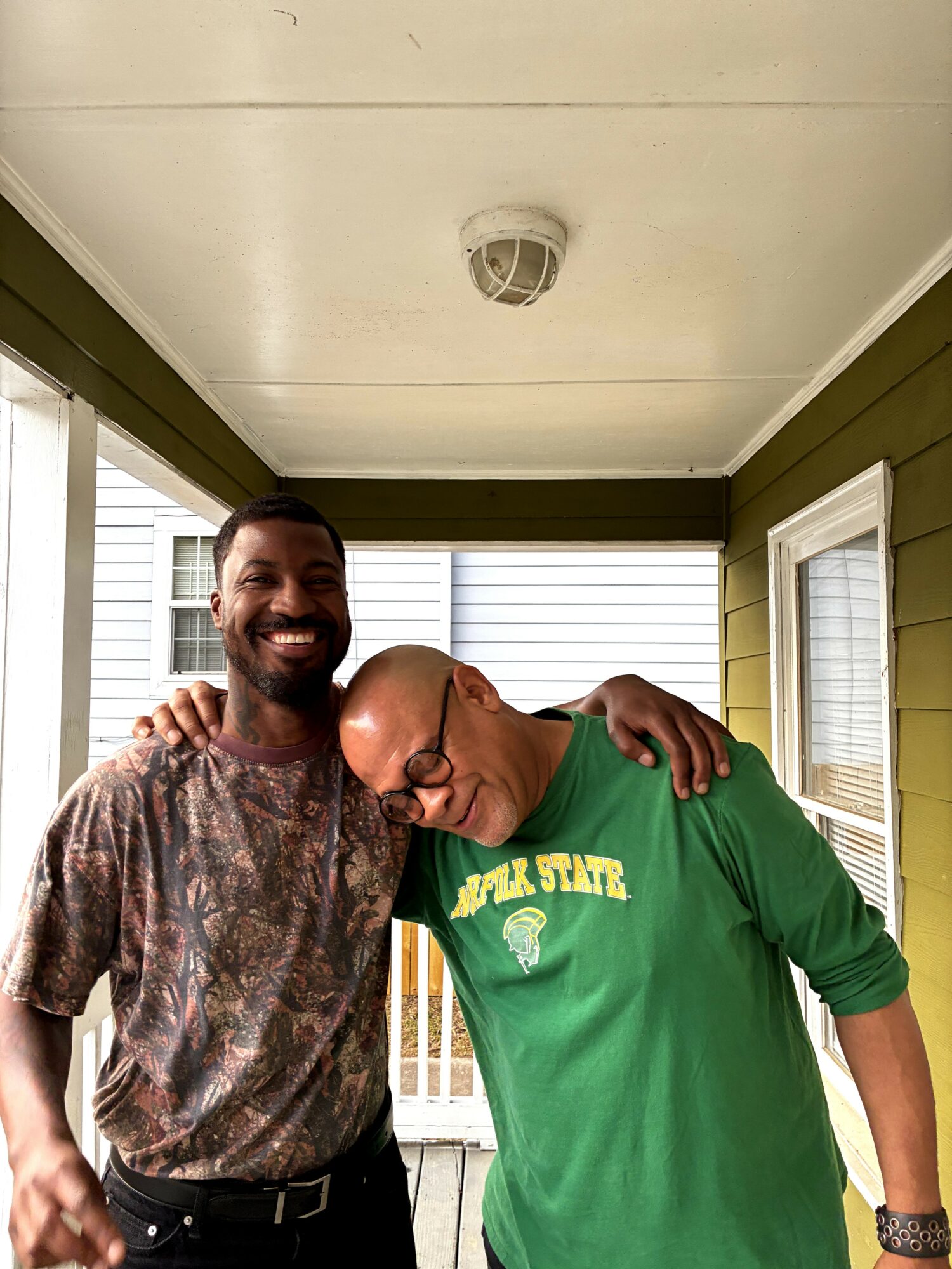Two men standing on a porch, smiling, with one resting his head on the other's shoulder, green and patterned shirts, outdoor setting.
