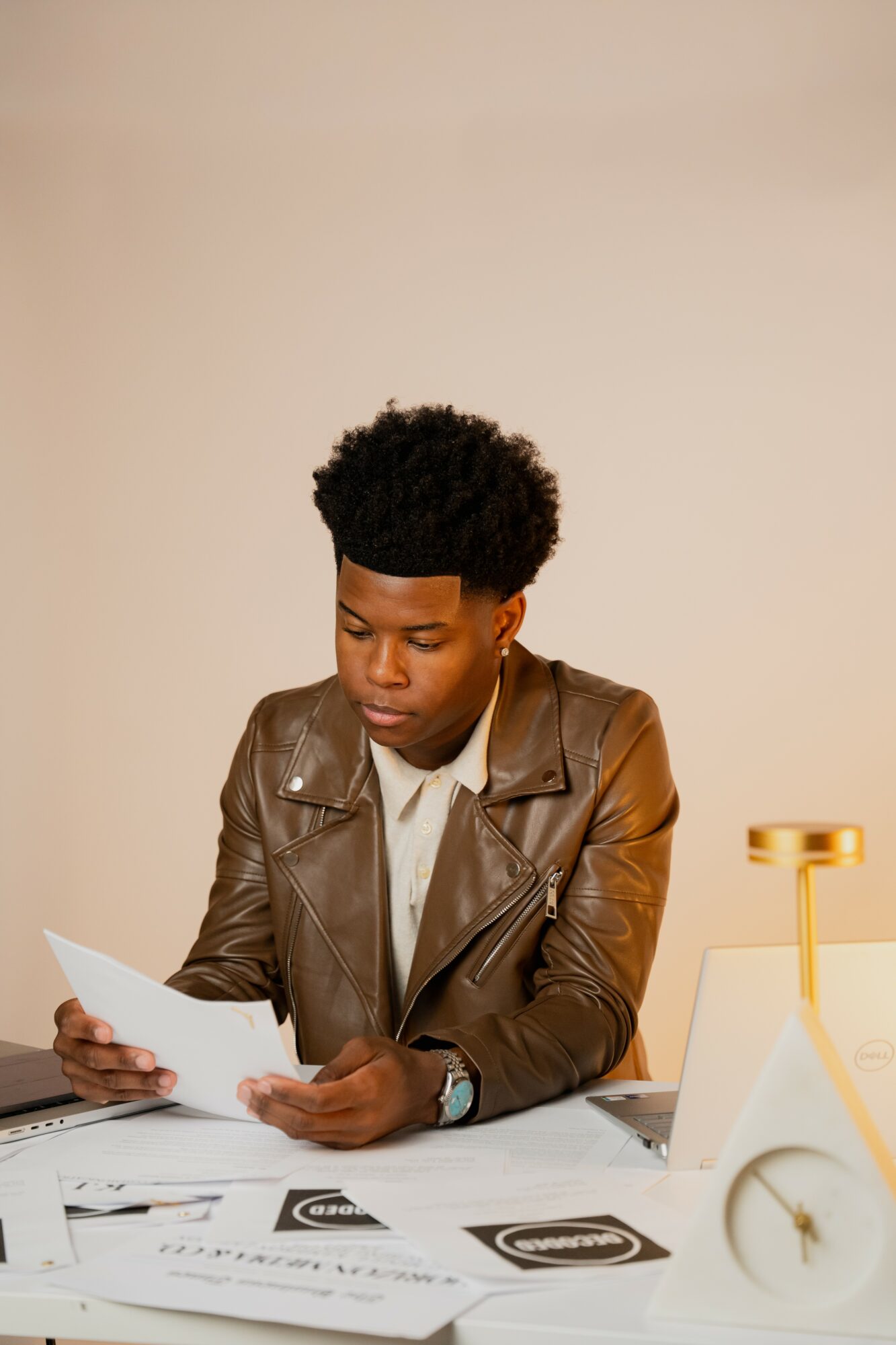 Young man with curly hair wearing a leather jacket, sitting at a desk, reading a paper, with a laptop and documents.