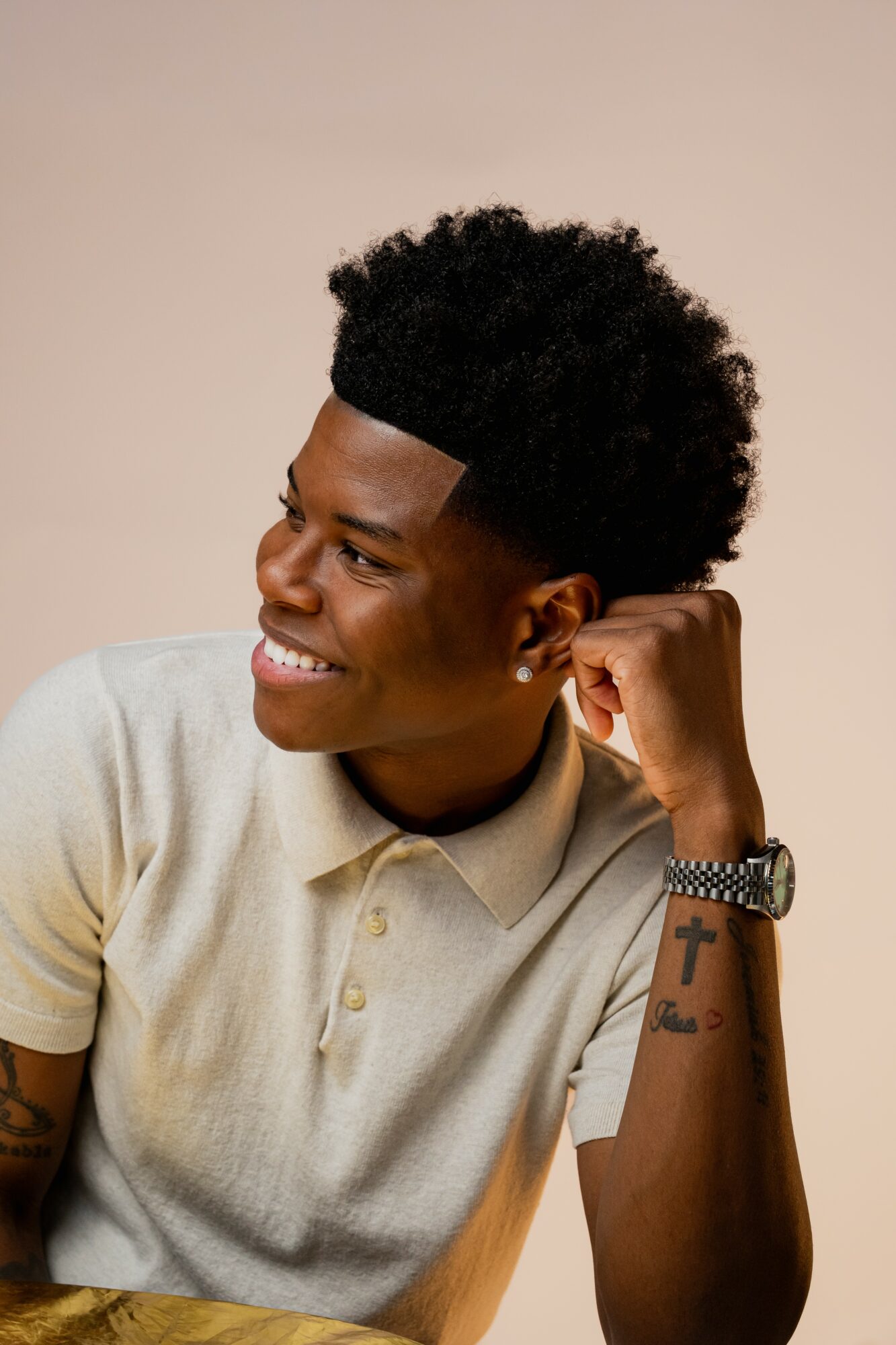Young person with short curly hair smiling, resting chin on hand, wearing a light-colored polo shirt and bracelet.