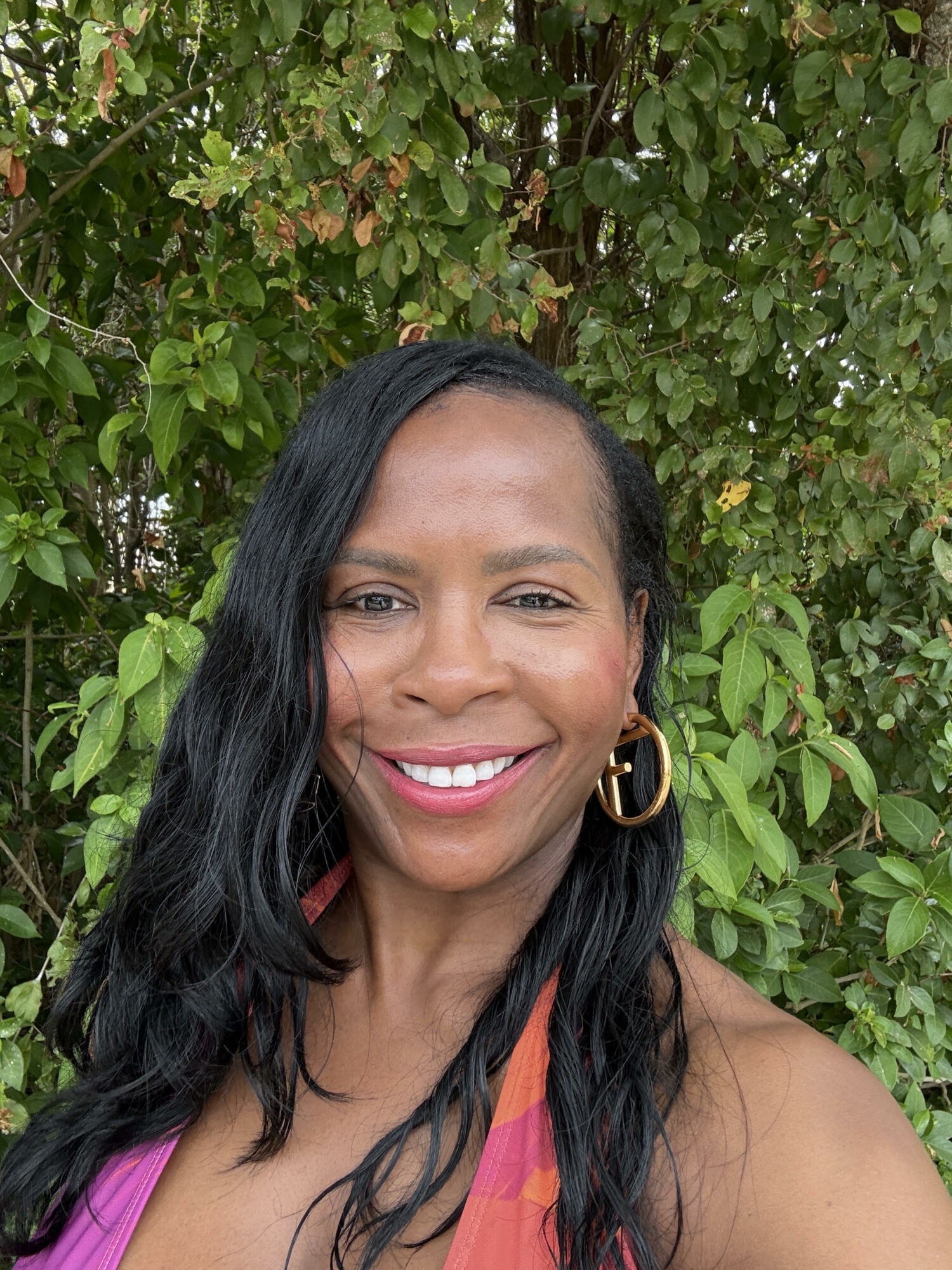 Smiling woman with long black hair, wearing gold hoop earrings, outdoors with green foliage background.