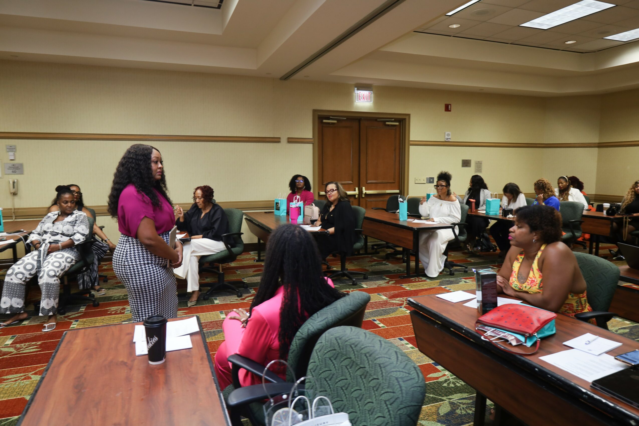 A woman stands and speaks to a seated group in a conference room with tables and chairs, some people taking notes.