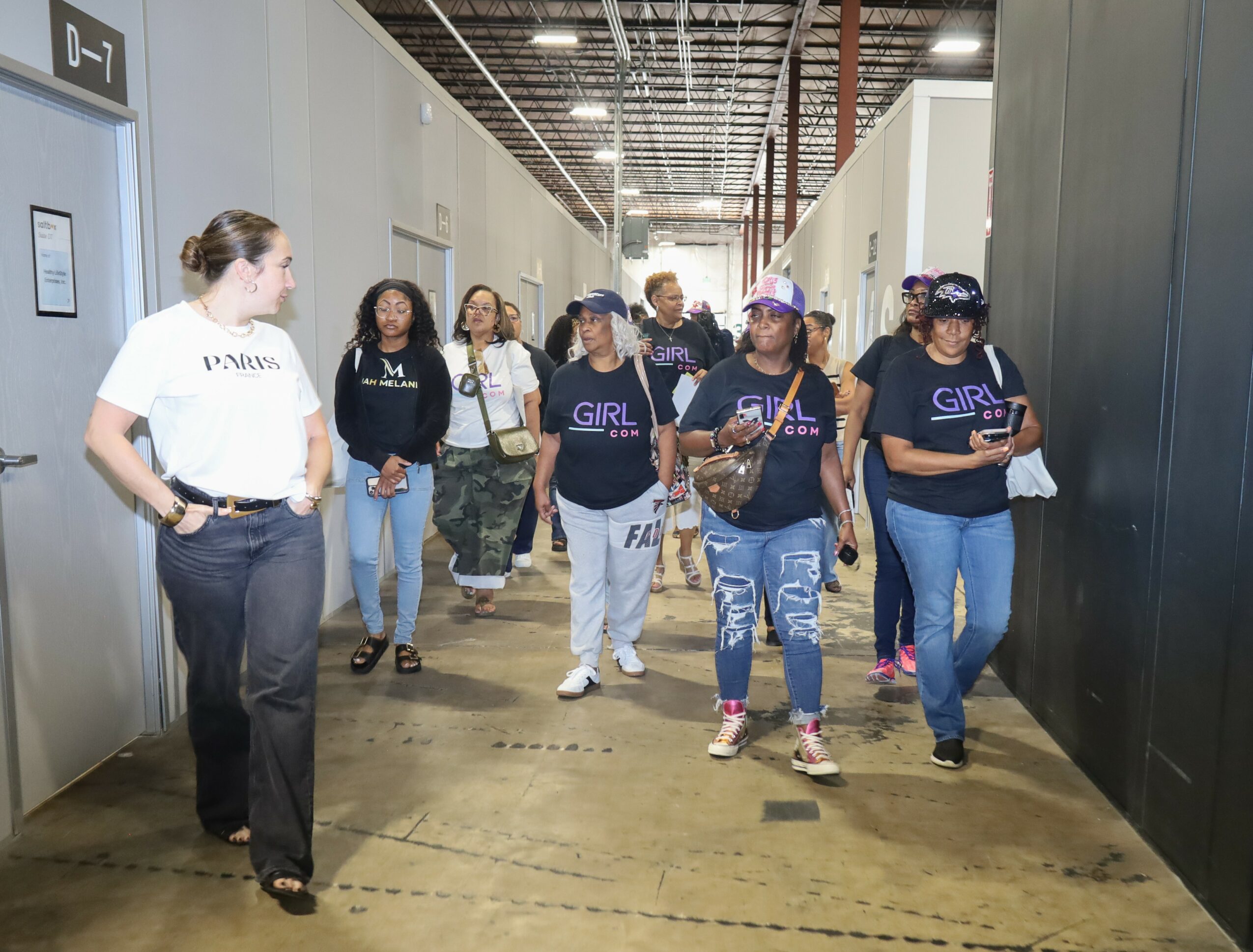 Group of women walking in a hallway, some wearing matching shirts, others with backpacks, in an industrial setting.