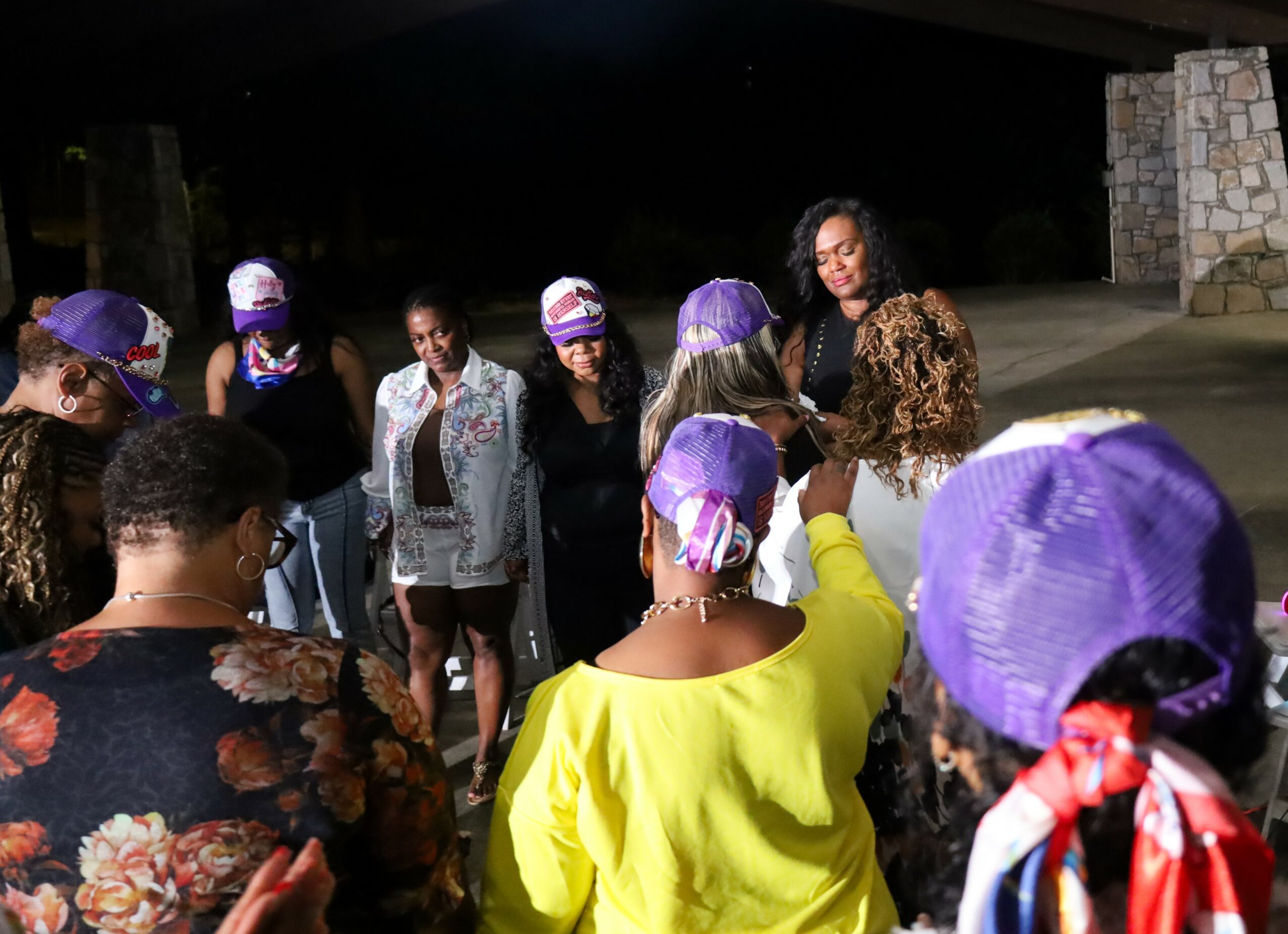 Group of people gathered outdoors at night, some wearing purple hats, listening to a woman speaking.