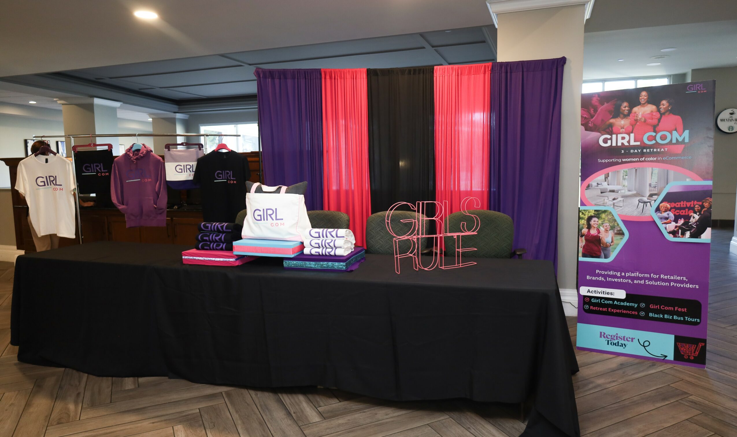 Table with purple and pink curtains, promotional materials, and a Girl.com banner at an indoor event.
