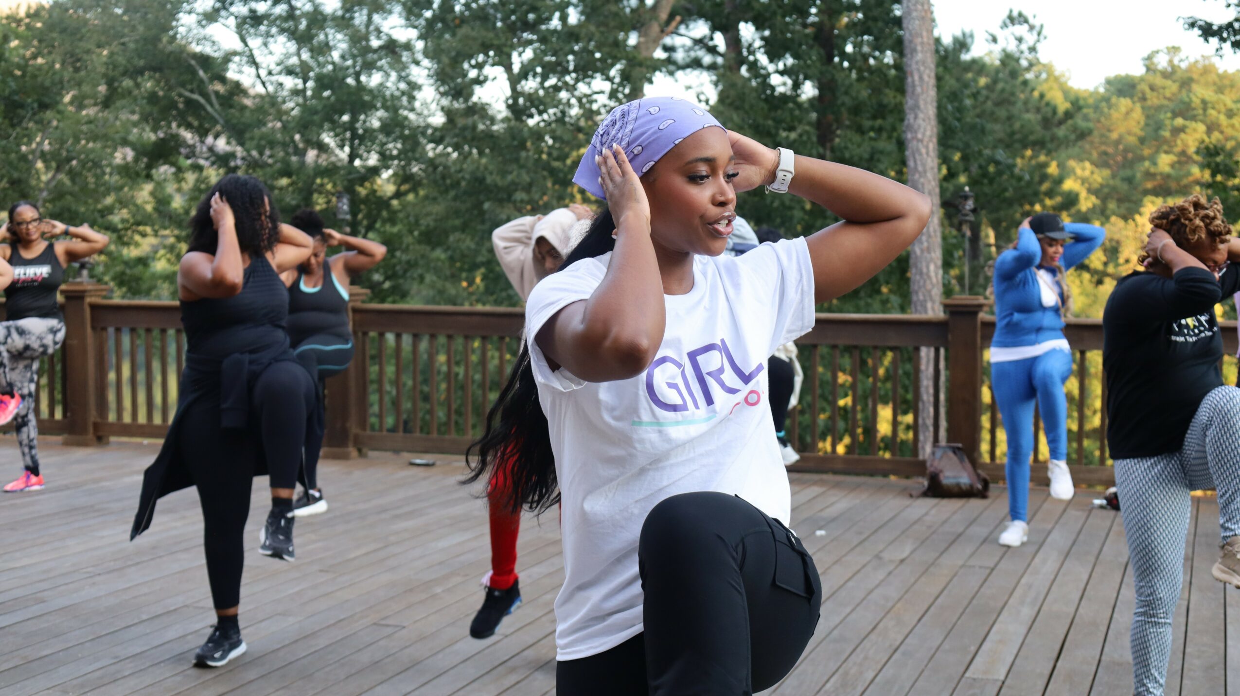 Group of people practicing dance outdoors on a wooden deck with trees in the background.